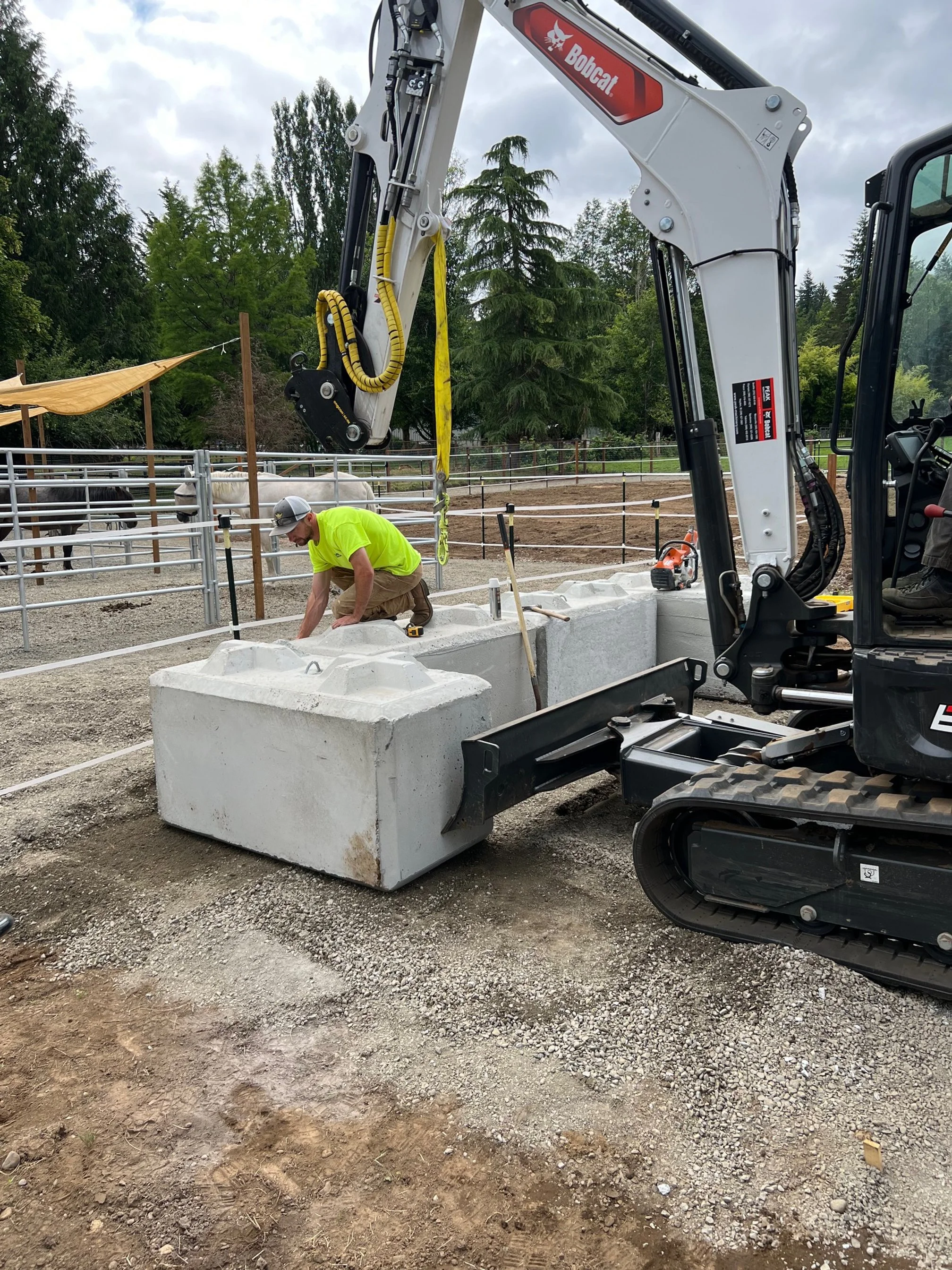 Construction worker crouching next to large concrete blocks in a partially dug outdoor area with a small excavator nearby and a fenced enclosure with horses and trees in the background.