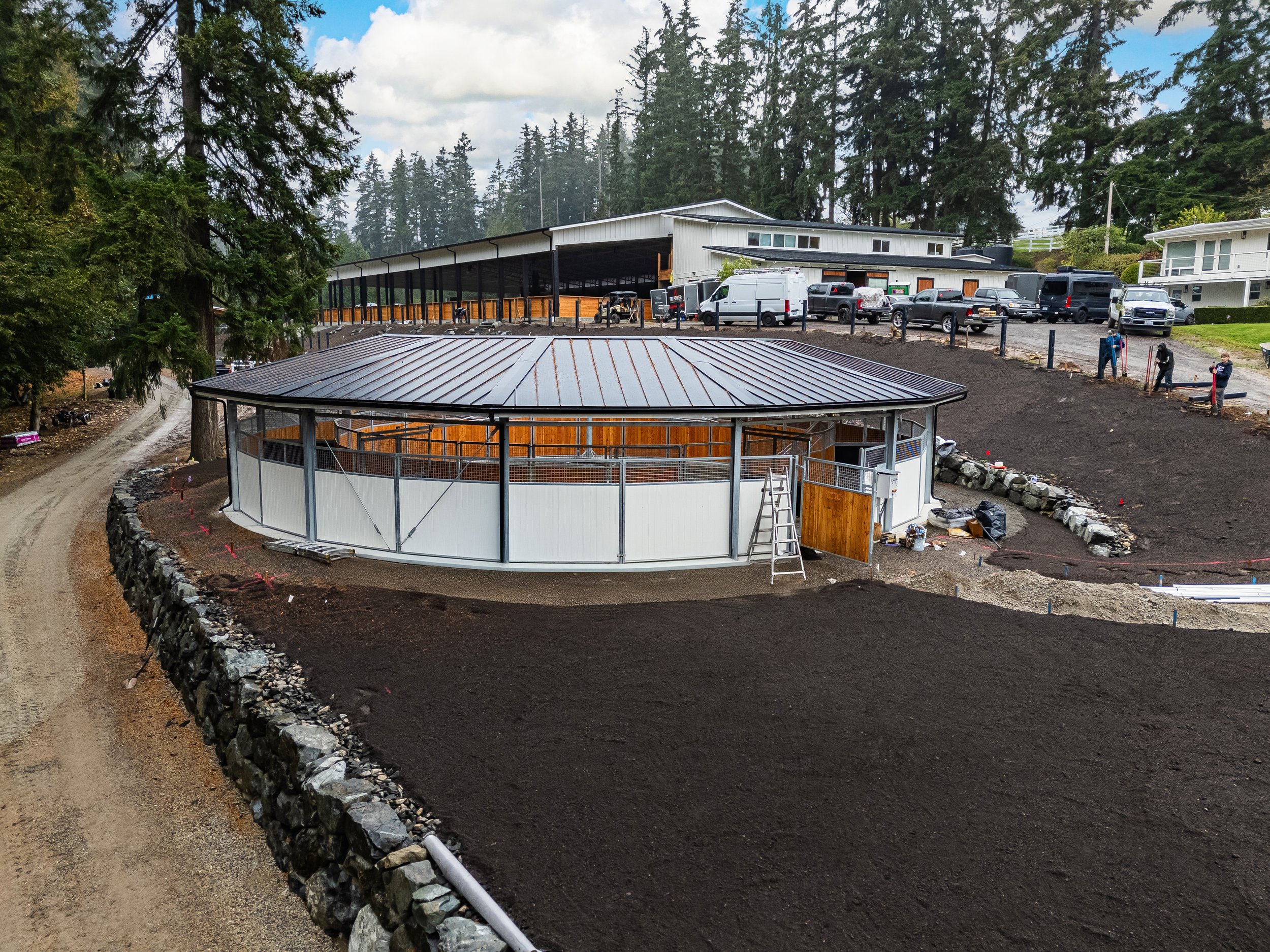 Construction site with a circular area surrounded by rocks, a slope with fresh soil, a large building with parking lot full of cars in the background, tall trees, and people working on the site.