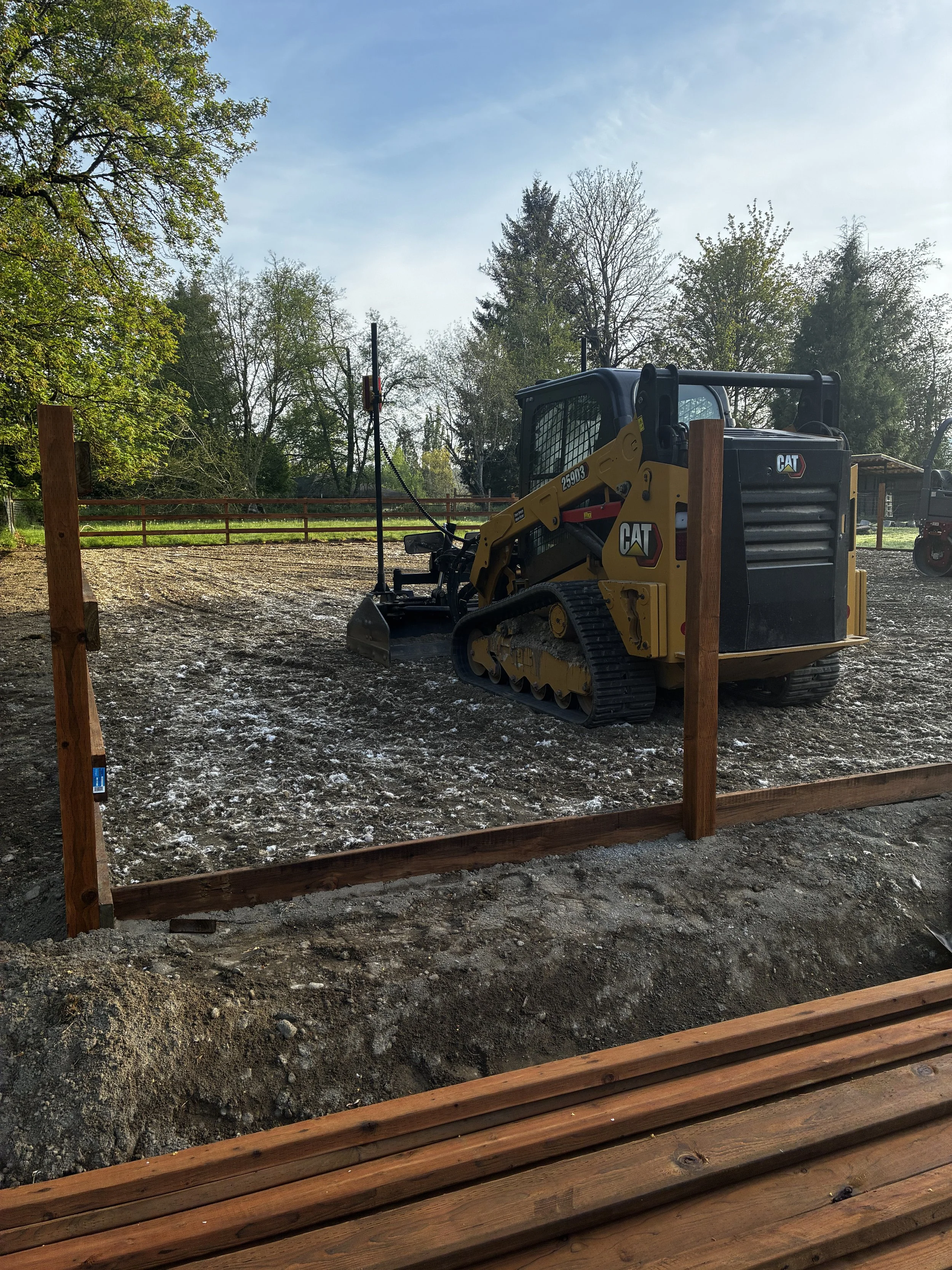 Construction site with a small Caterpillar bulldozer, wooden framing, and trees in the background under a partly cloudy sky.