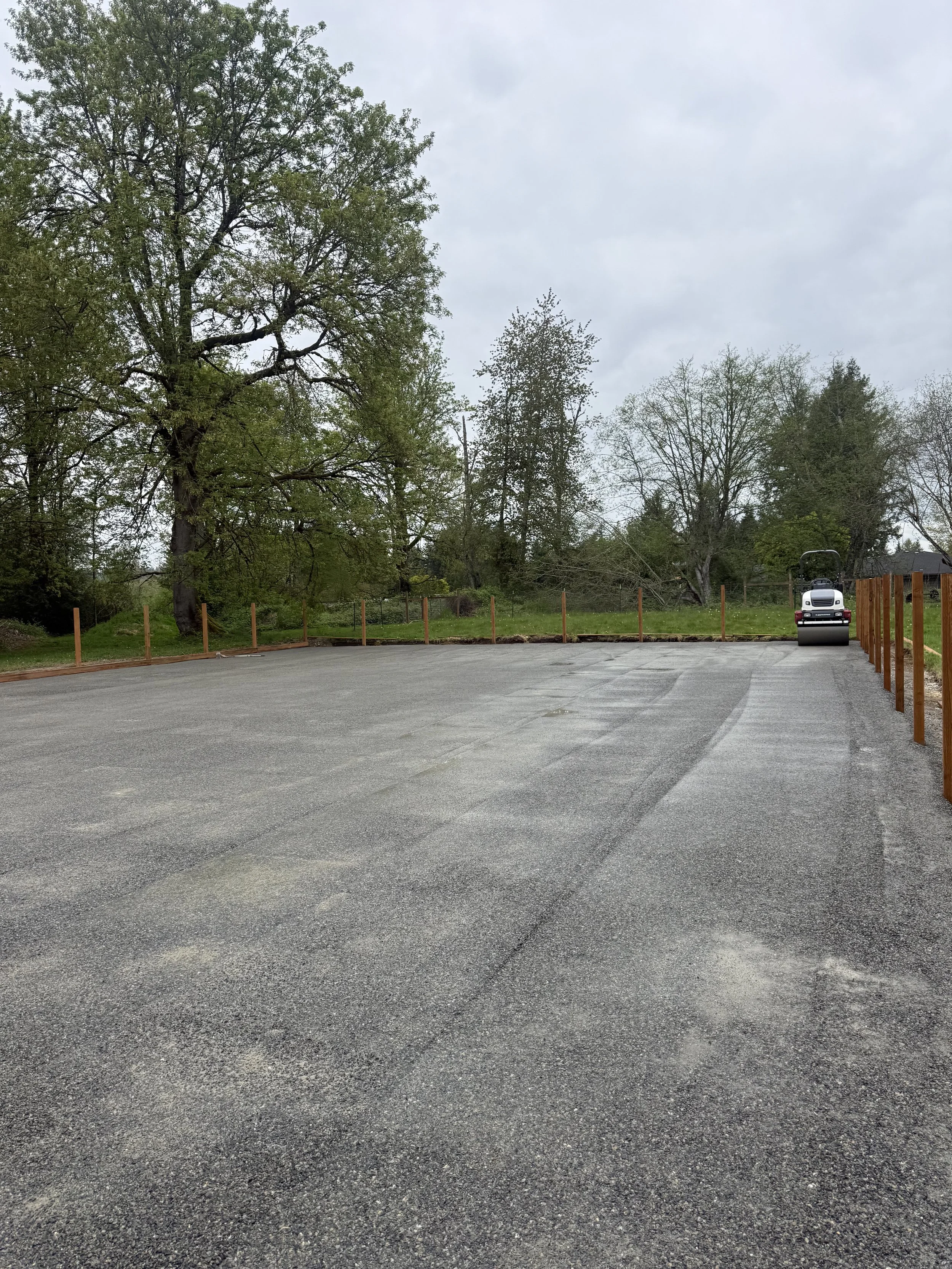 Empty paved parking lot with a view of trees and overcast sky in the background, and a white utility truck parked far in the distance.