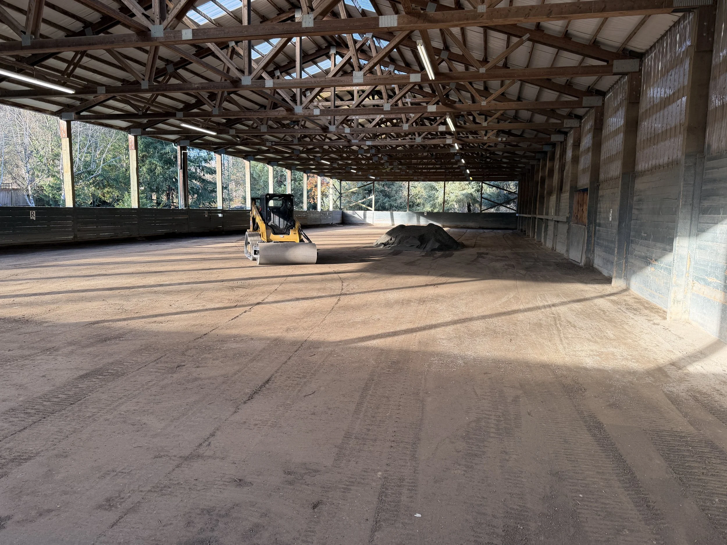Construction site with a roller compactor on a dirt floor inside a large open wooden structure with a peaked roof and overhead lighting, surrounded by trees outside.