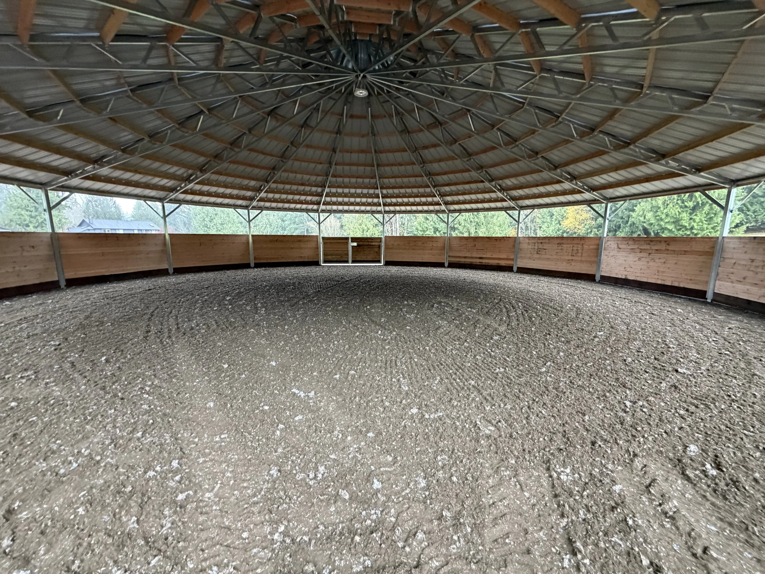 Indoor riding arena with a dirt floor, wooden fencing, and a roof supported by metal beams.