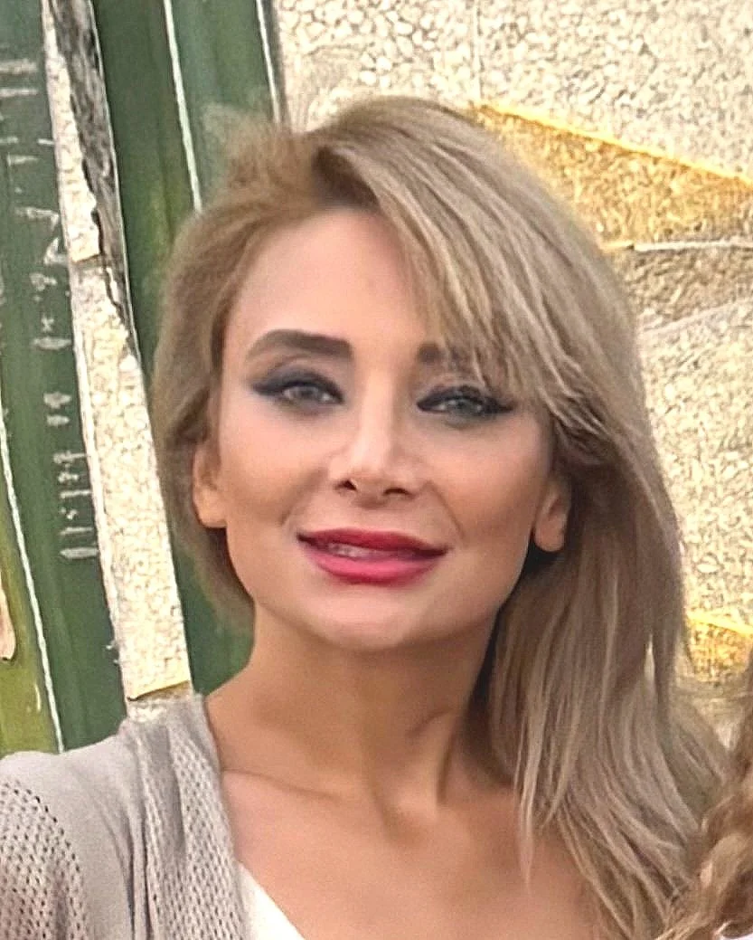 A woman with blonde hair styled in loose waves, wearing makeup with dark eye makeup and pink lipstick, smiling at the camera. Background appears to be an outdoor setting with a green structure and a stone or concrete surface.