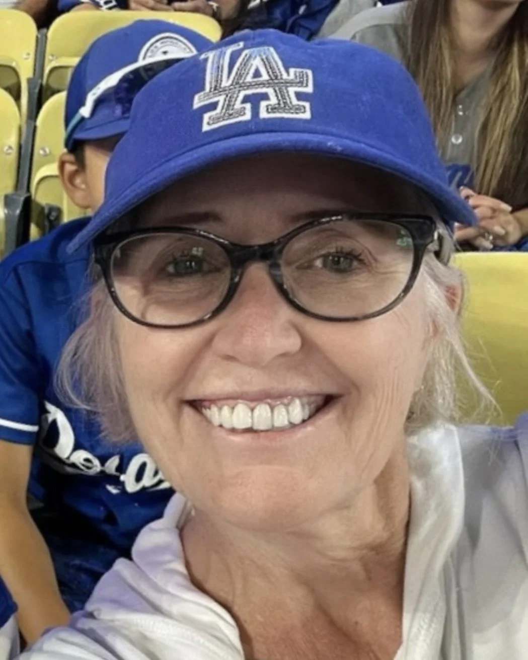 Smiling woman wearing glasses and a blue Los Angeles Dodgers cap taking a selfie at a sports event with people seated in yellow chairs behind her.