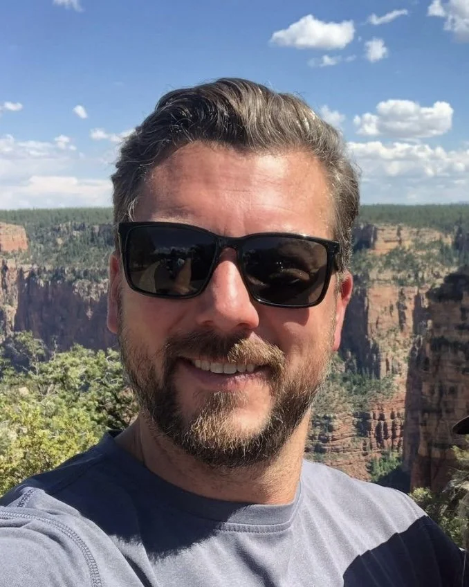 A man with sunglasses and a beard taking a selfie at the Grand Canyon with a clear blue sky and clouds overhead.