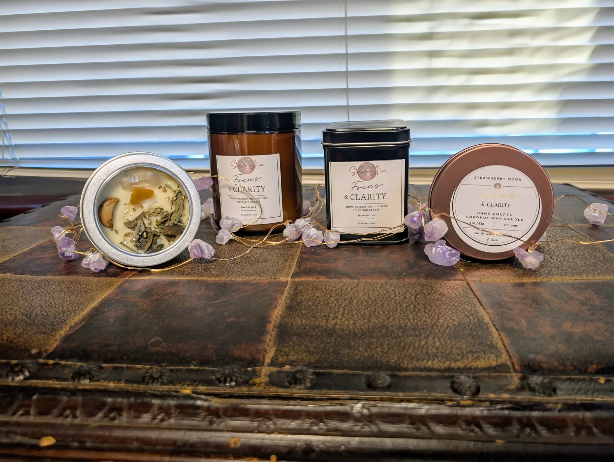 Collection of scented candles with labels and decorative stones on a wooden surface with a window in the background.