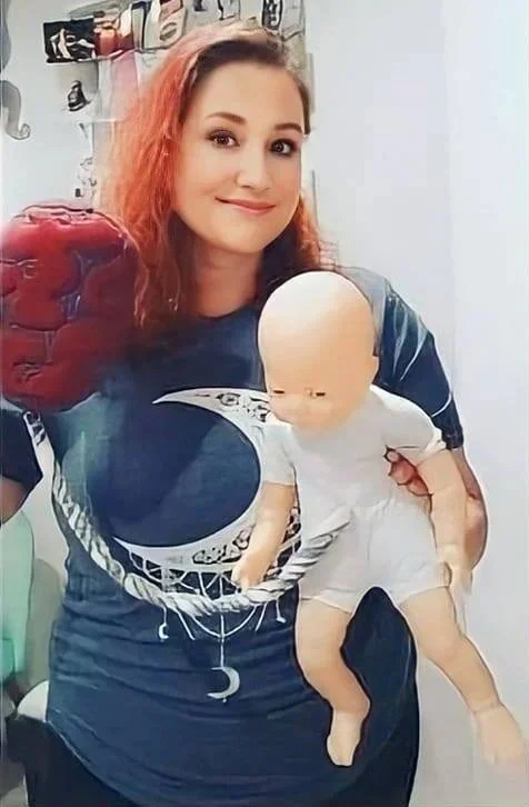 A woman with shoulder-length curly red hair smiling at the camera, holding a doll that resembles a human baby.