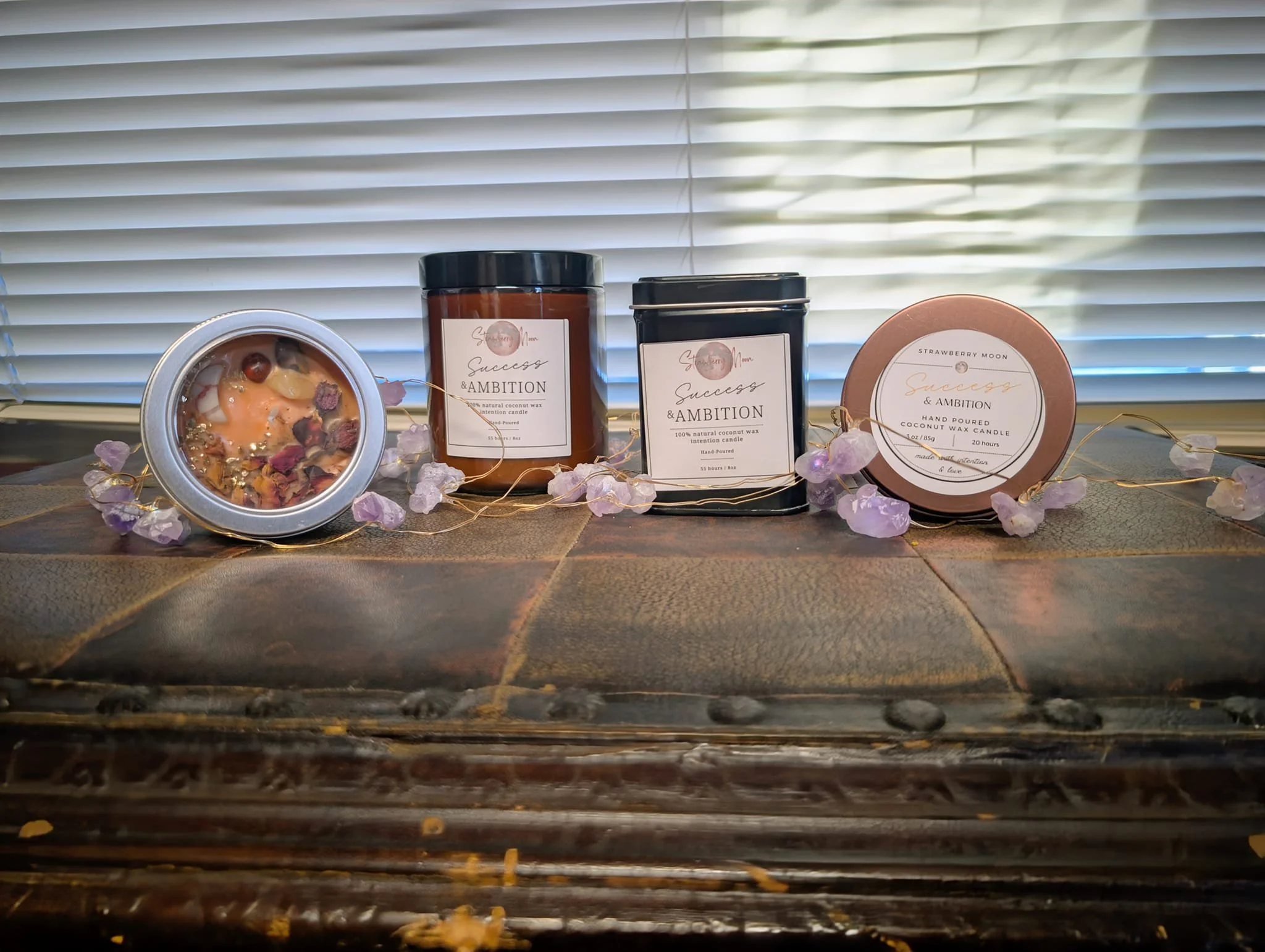 Collection of scented candles with labels and decorative stones on a wooden surface with a window in the background.