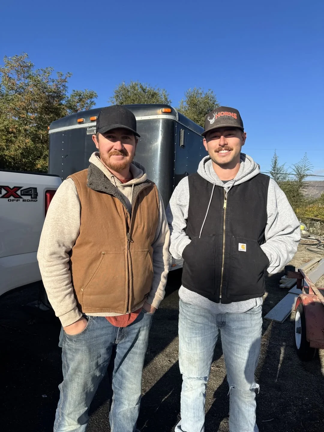 Two men standing outdoors in front of a trailer and pickup truck, dressed in casual work clothing, smiling at the camera under a clear blue sky.