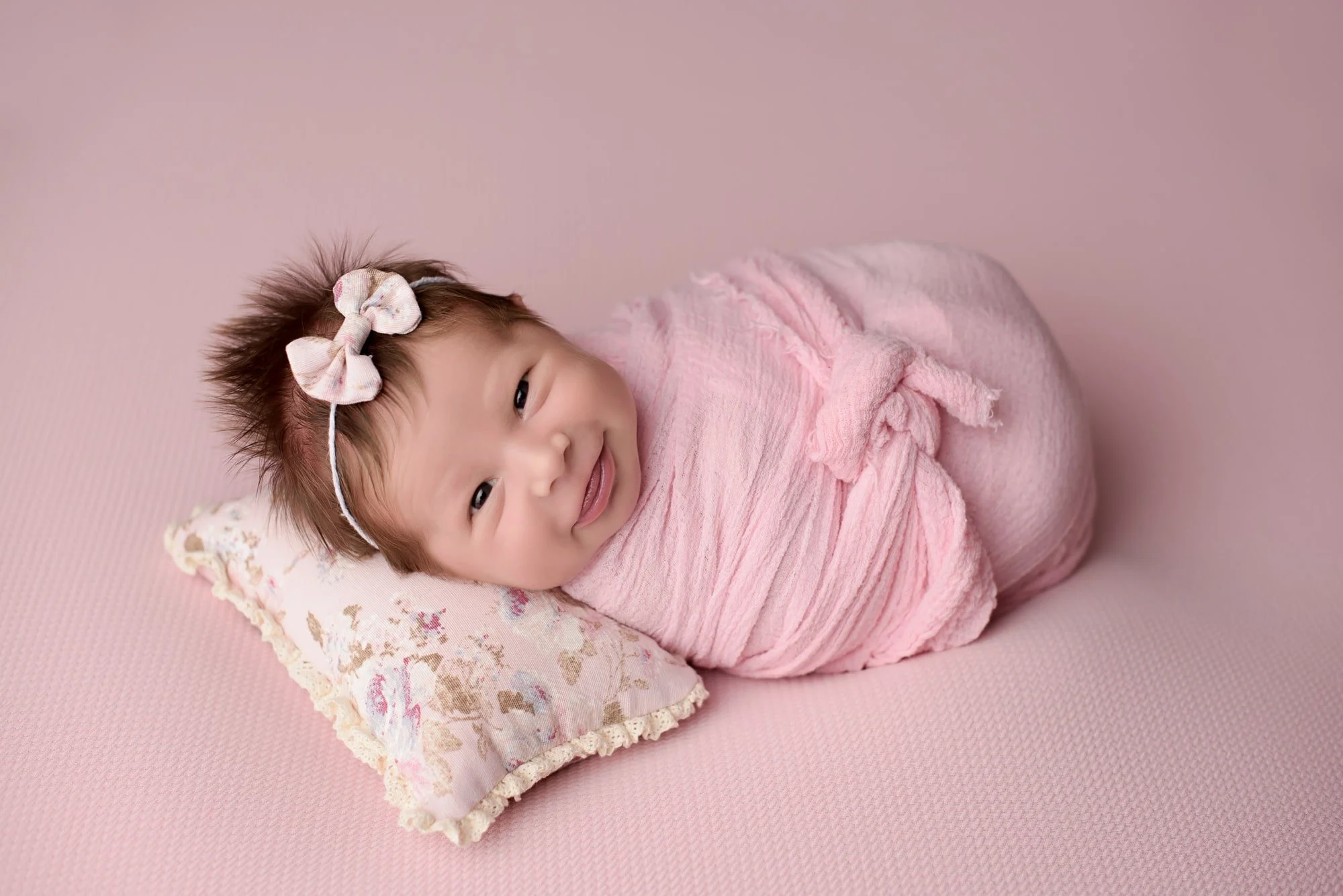 newborn infant wrapped in pink blanket laying on a floral pillow with a pink background