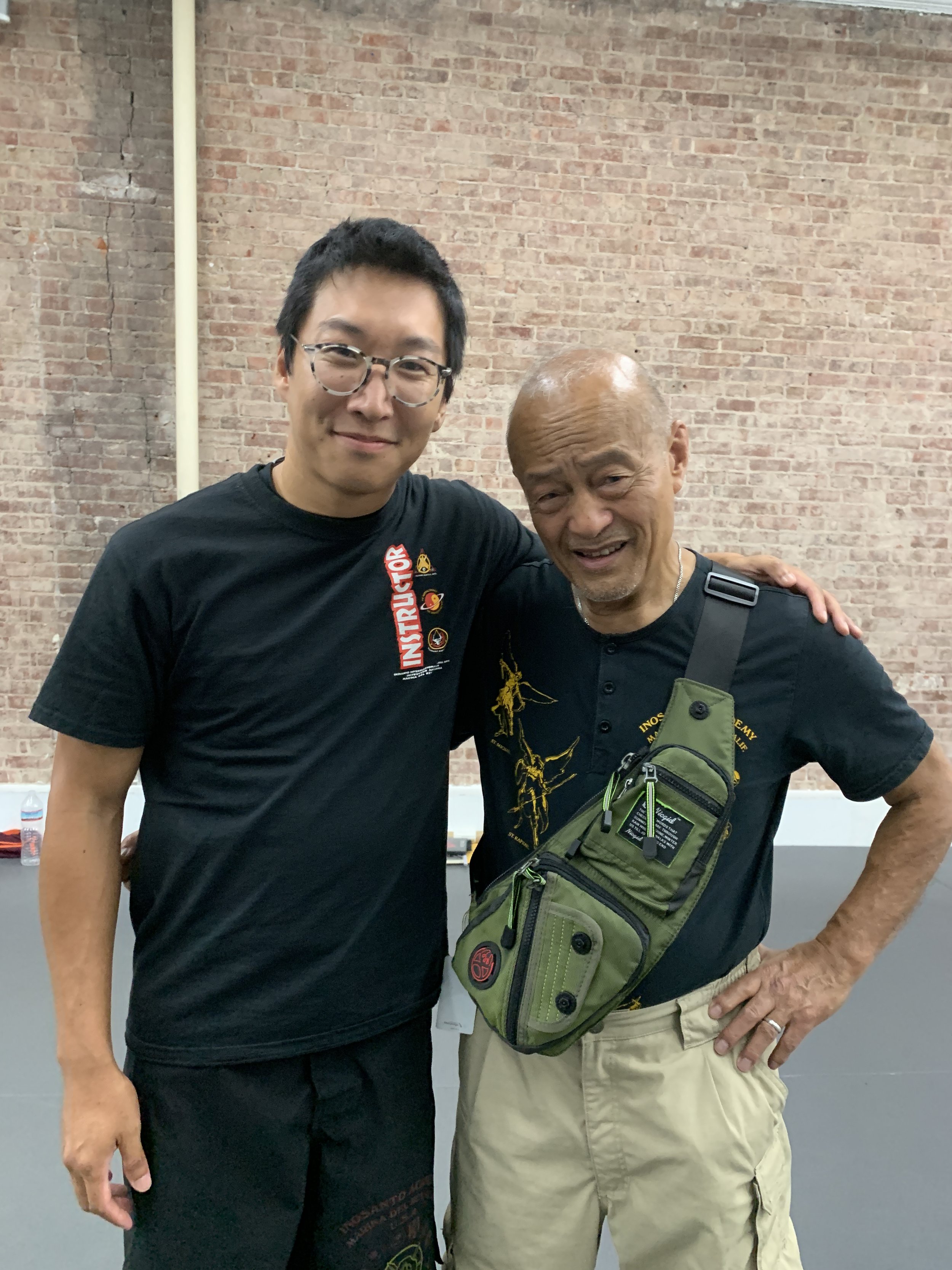 Two men standing together, smiling, with their arms around each other in front of a brick wall. One is young with black hair and glasses, wearing a black T-shirt. The other is older, bald, with a goatee, wearing a dark T-shirt, khaki pants, and a green crossbody bag.