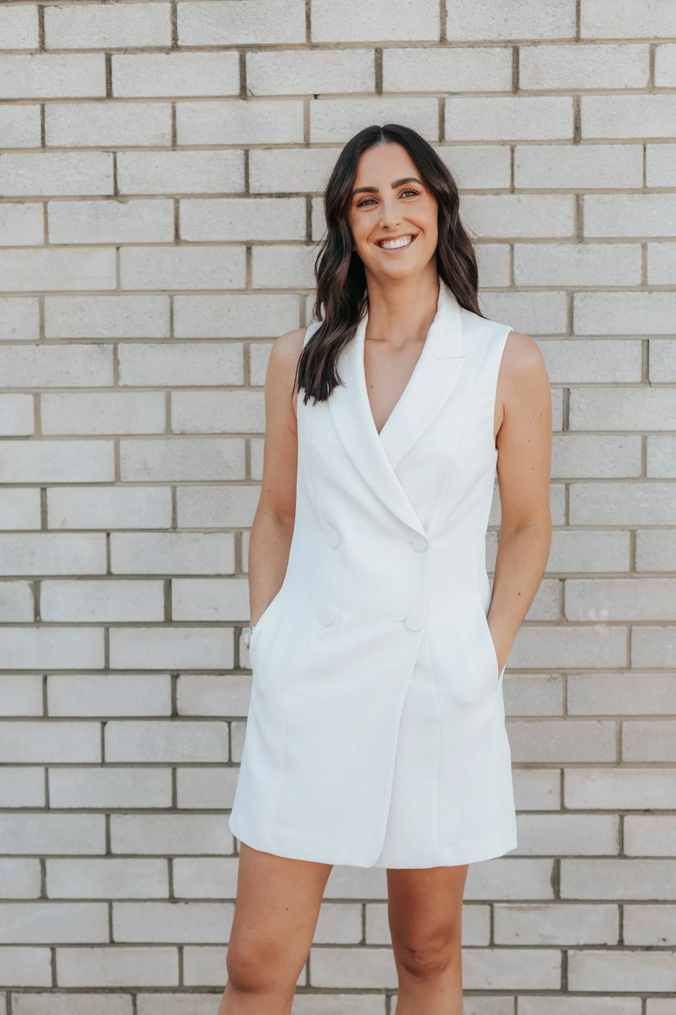 Woman in a white sleeveless dress standing against a brick wall.