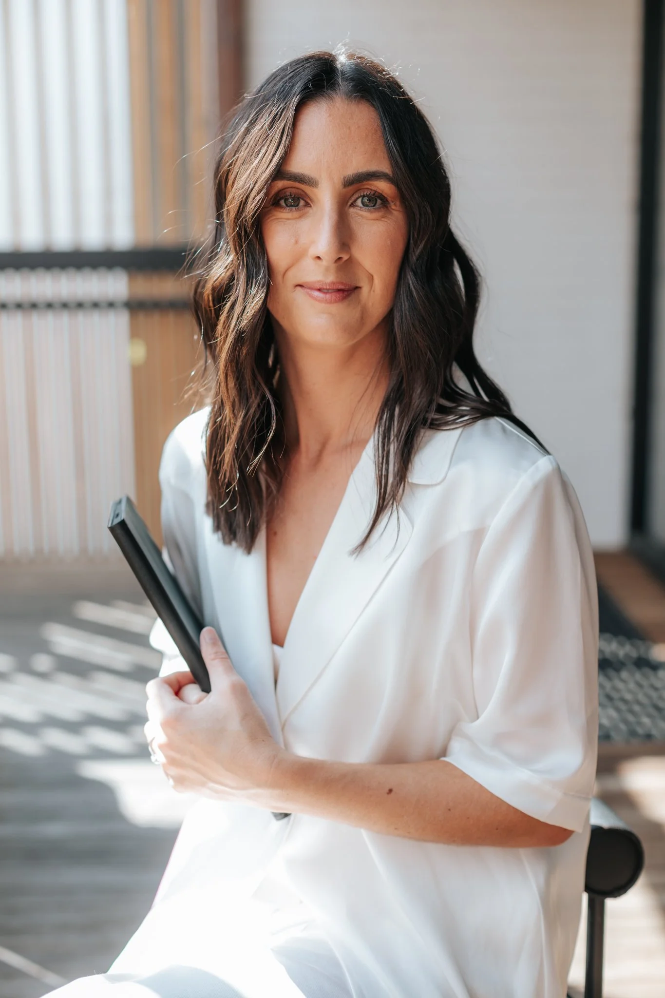 Woman with long dark hair in a white blouse holding a book, sitting in a sunlit room.