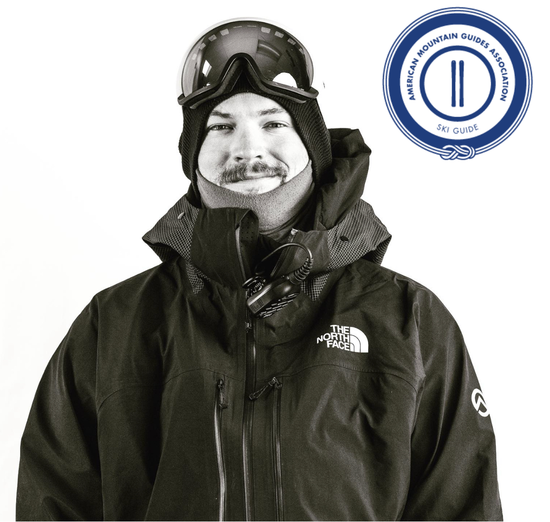 A man dressed in winter skiing gear including a black North Face jacket, ski goggles on his forehead, and a black beanie, standing in front of a white background with an American Mountain Guides Association logo in the top right corner.