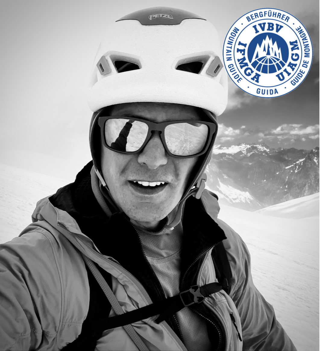 A man in mountain gear, wearing a helmet and sunglasses, taking a selfie on a snowy mountain with mountains and clouds in the background. There is a round blue and white logo in the top right corner with text in multiple languages and mountain icons.