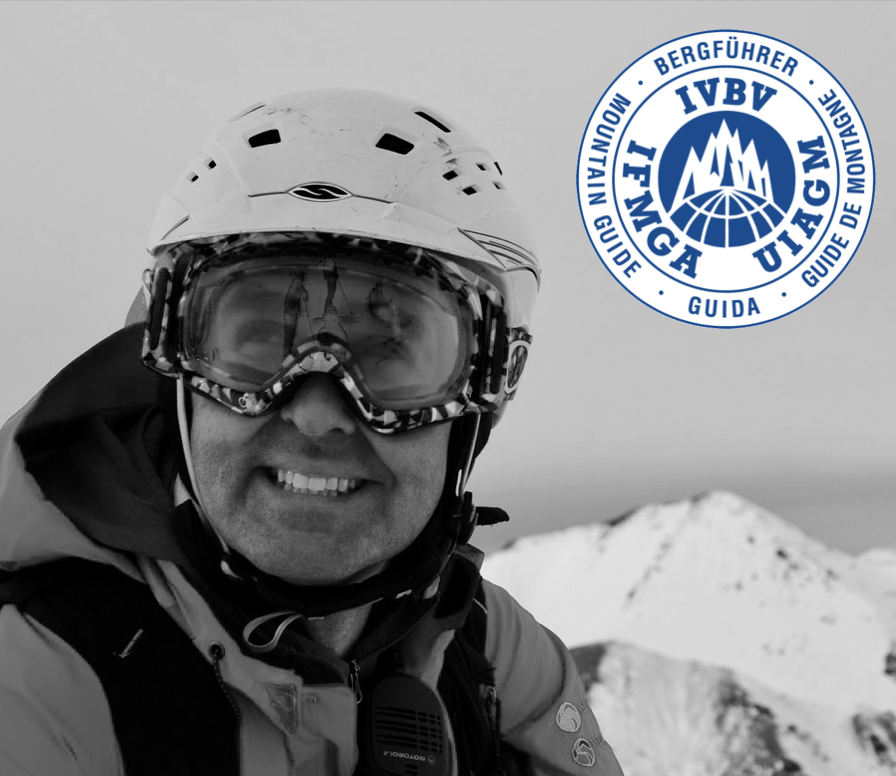 A man wearing a helmet and ski goggles smiling in front of snowy mountain peaks, with a logo in the top right corner that includes mountain and globe graphics and text in German and English.