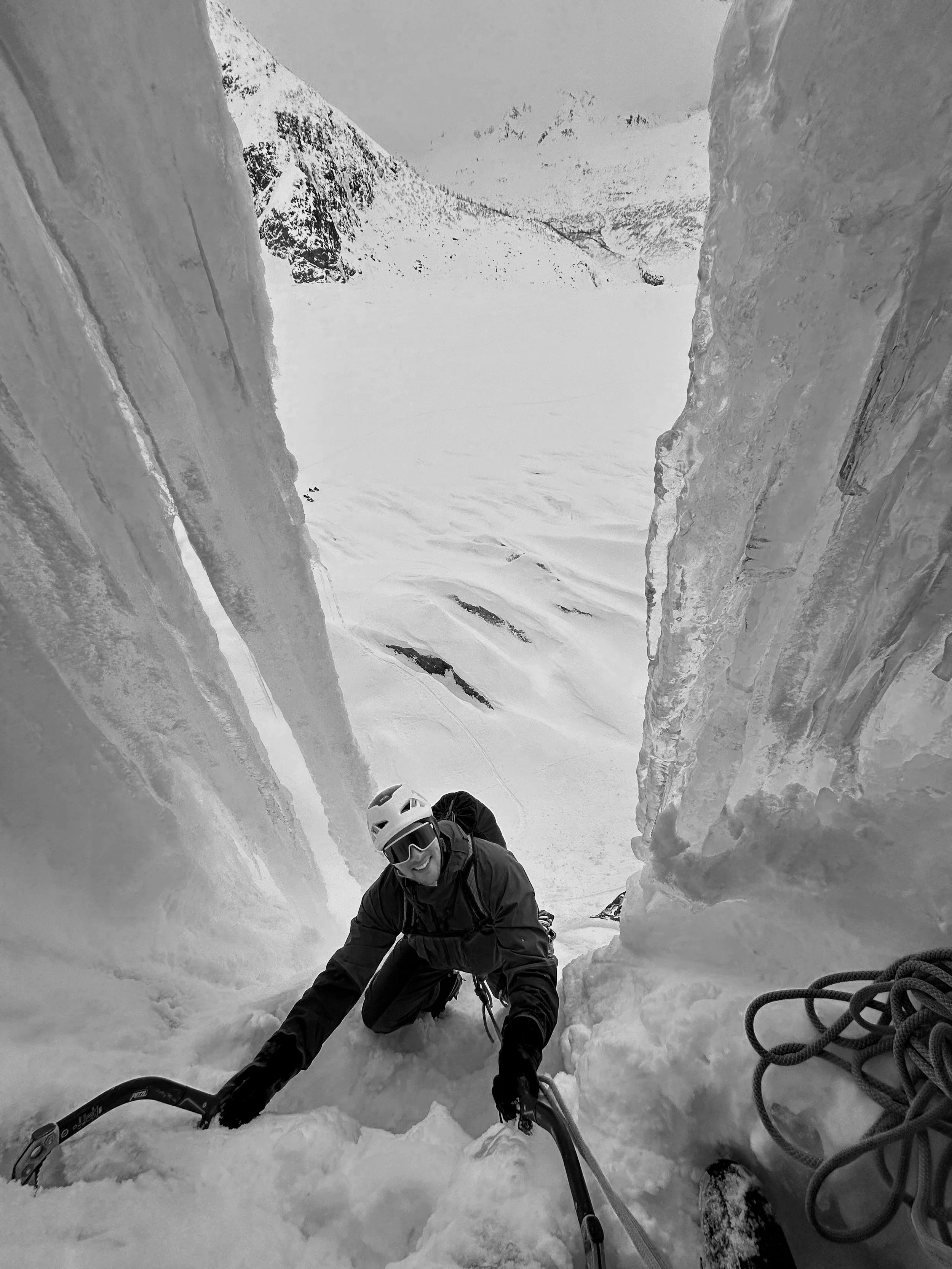 A climber in a helmet and ski gear ascending a narrow ice crevasse with snow-covered mountains in the background.