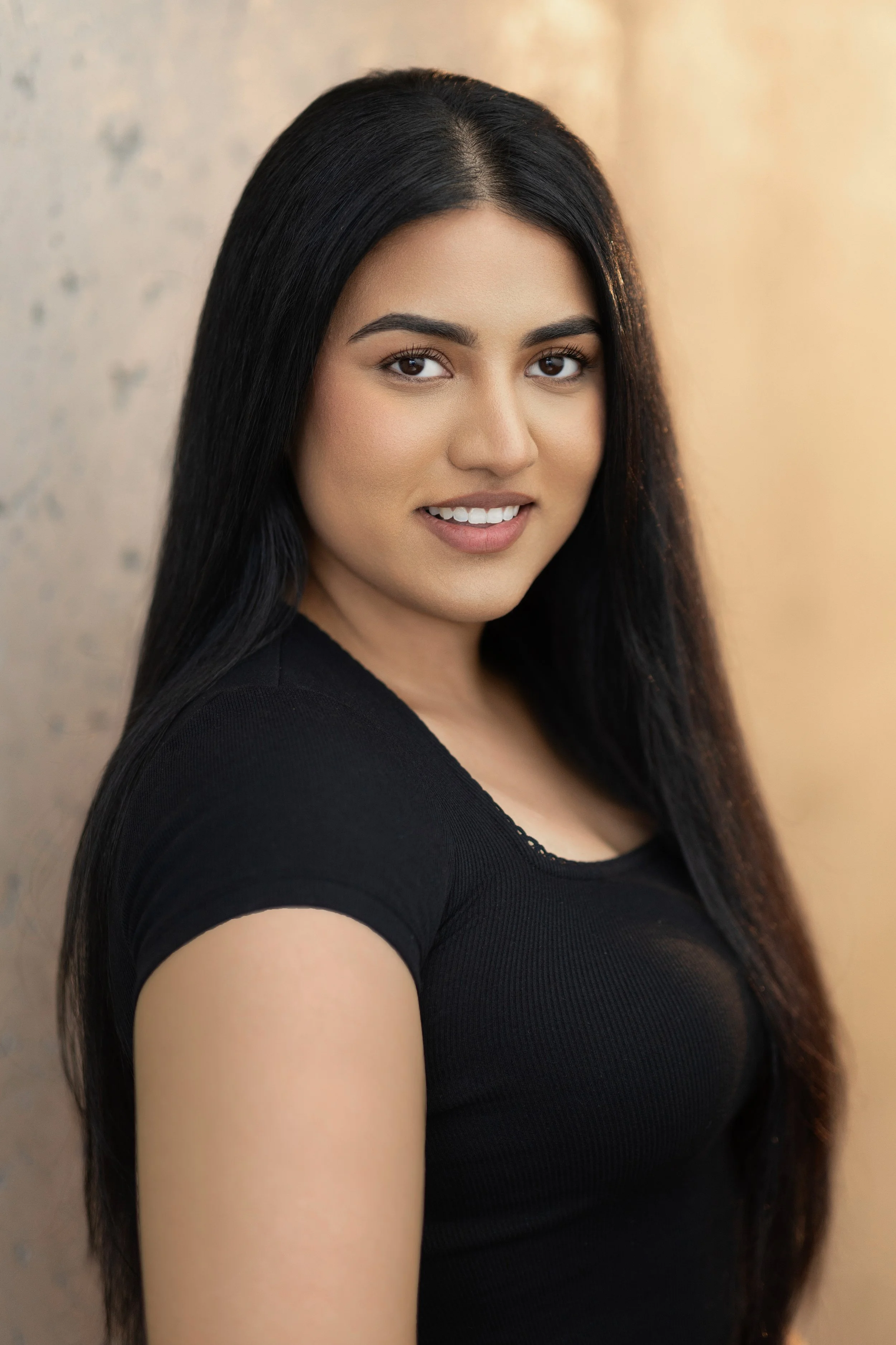 A young woman with long, straight black hair, wearing a black top, smiling and looking at the camera against a soft, blurred background.