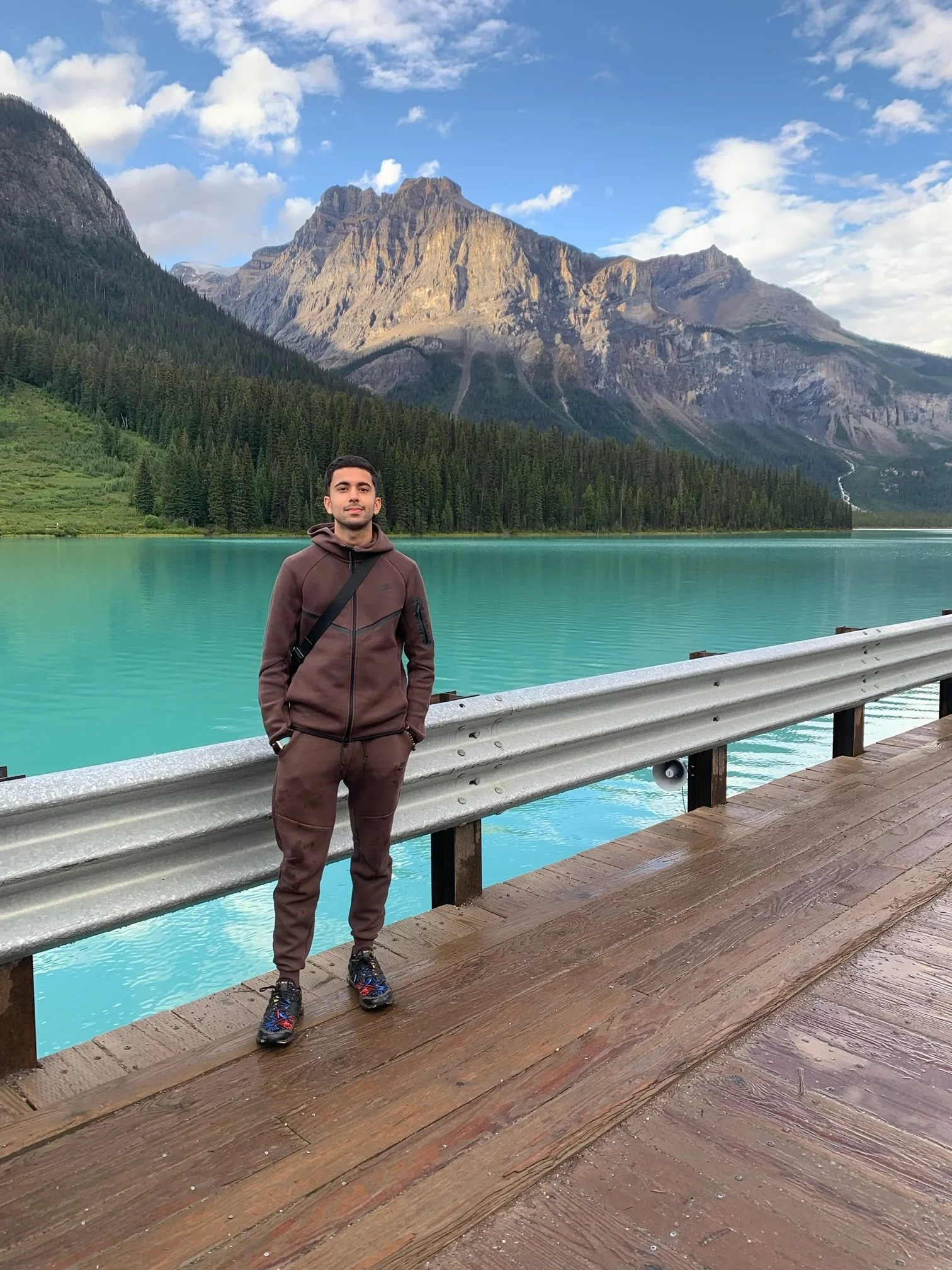 A man in a brown jacket and pants standing on a wooden deck by a turquoise lake with mountains and pine trees in the background under a partly cloudy sky.