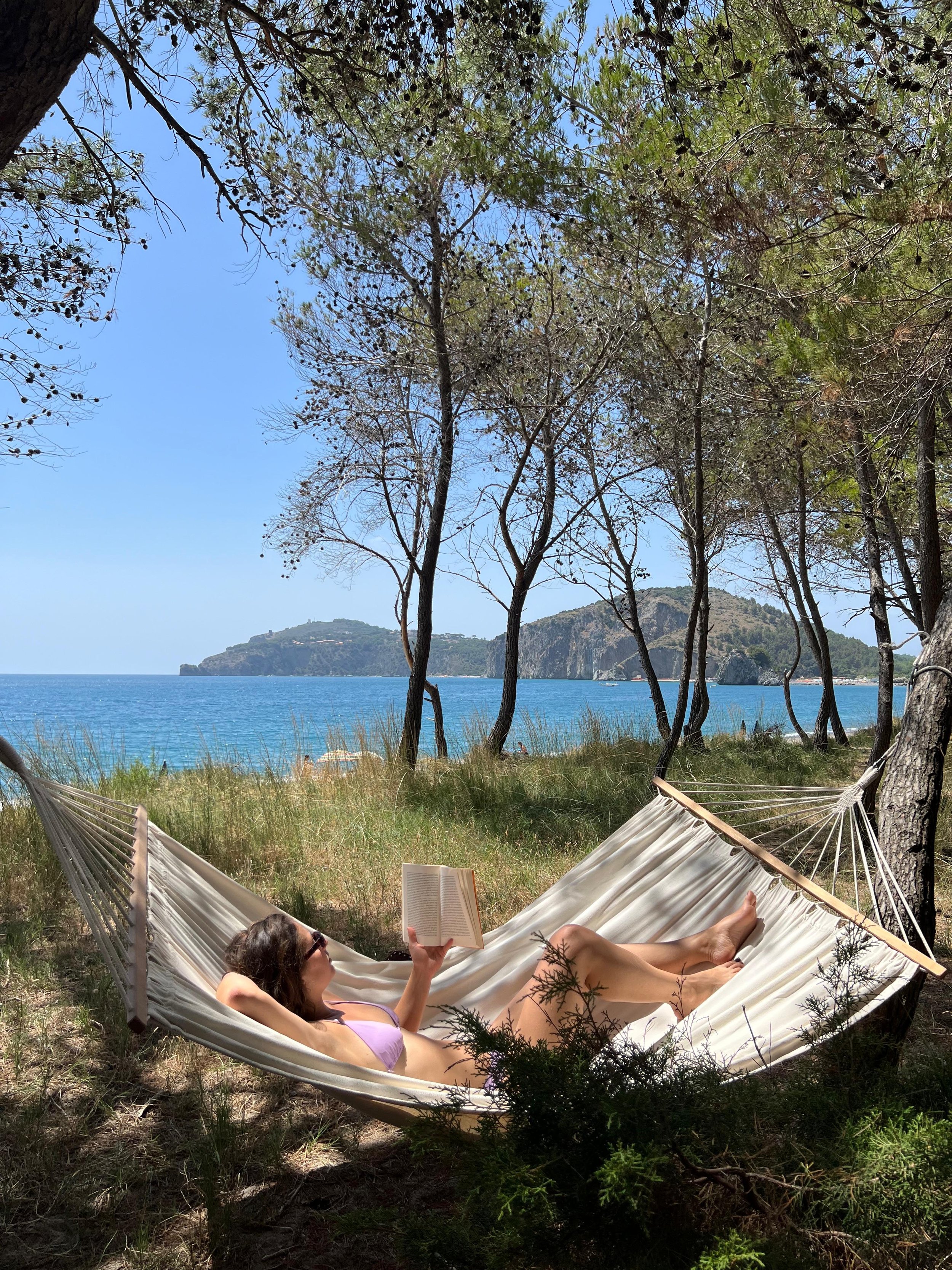 Una persona sdraiata su un'amaca tra gli alberi, vicino al mare, leggendo un libro. Il cielo è sereno e c'è una vista sulla costa e il mare in lontananza.