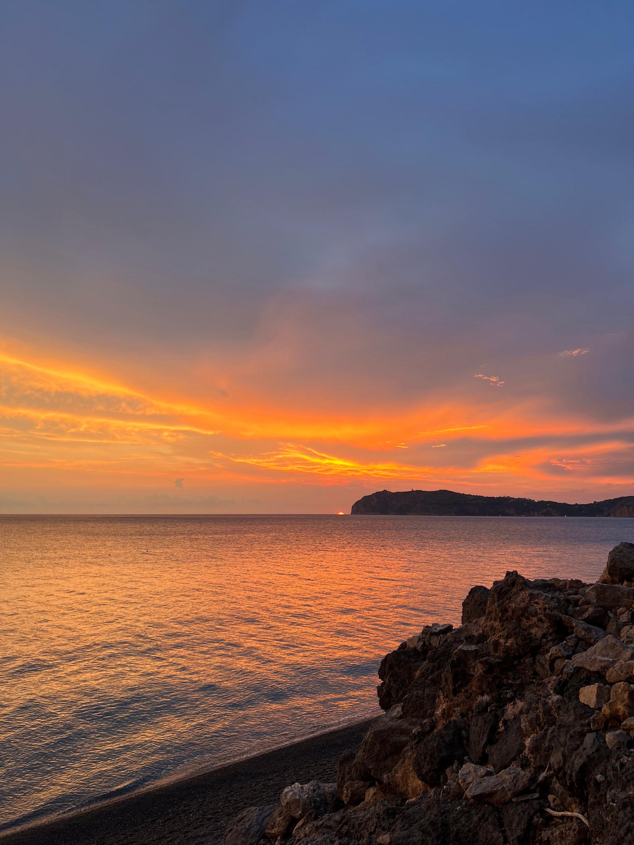 Tramonto sul mare con cielo arancione e blu, scogli in primo piano e collina all'orizzonte.