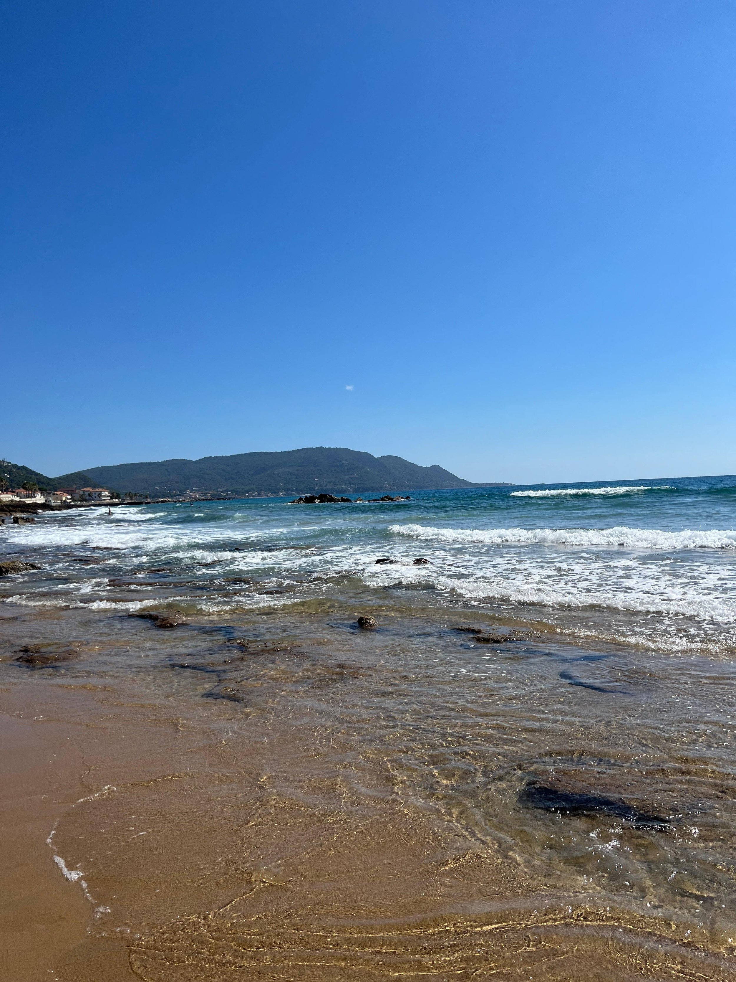 Spiaggia con sabbia e acqua cristallina, onde, cielo azzurro, colline sullo sfondo.