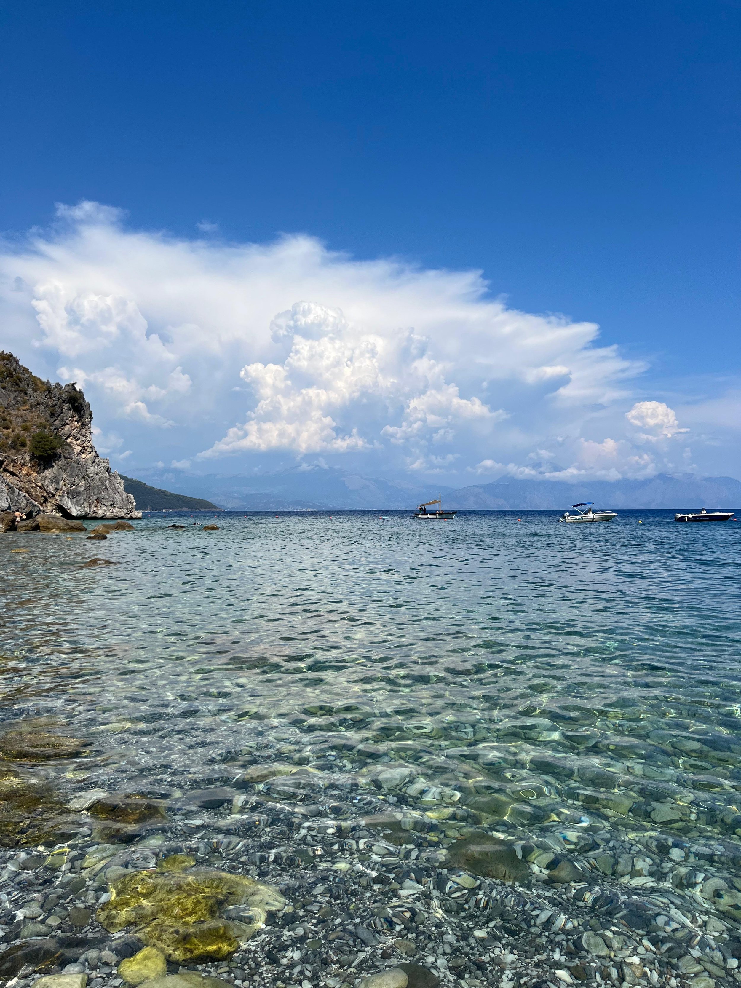 Spiaggia rocciosa con mare trasparente e cielo azzurro, alcune barche sullo sfondo.