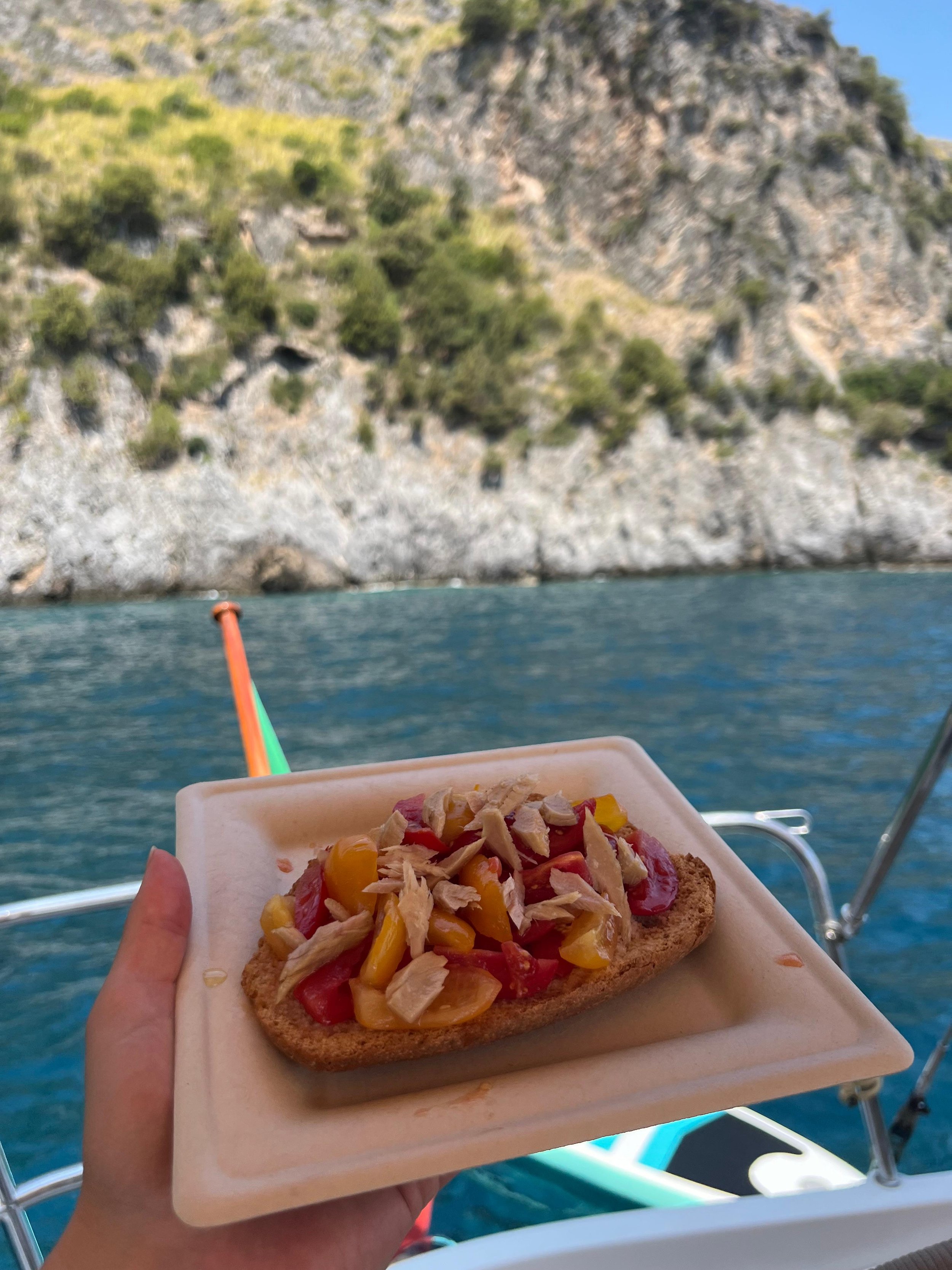 Persona tiene un piatto con una bruschetta, decorata con pomodori e tonno, su uno sfondo di mare e scogliere verdi.
