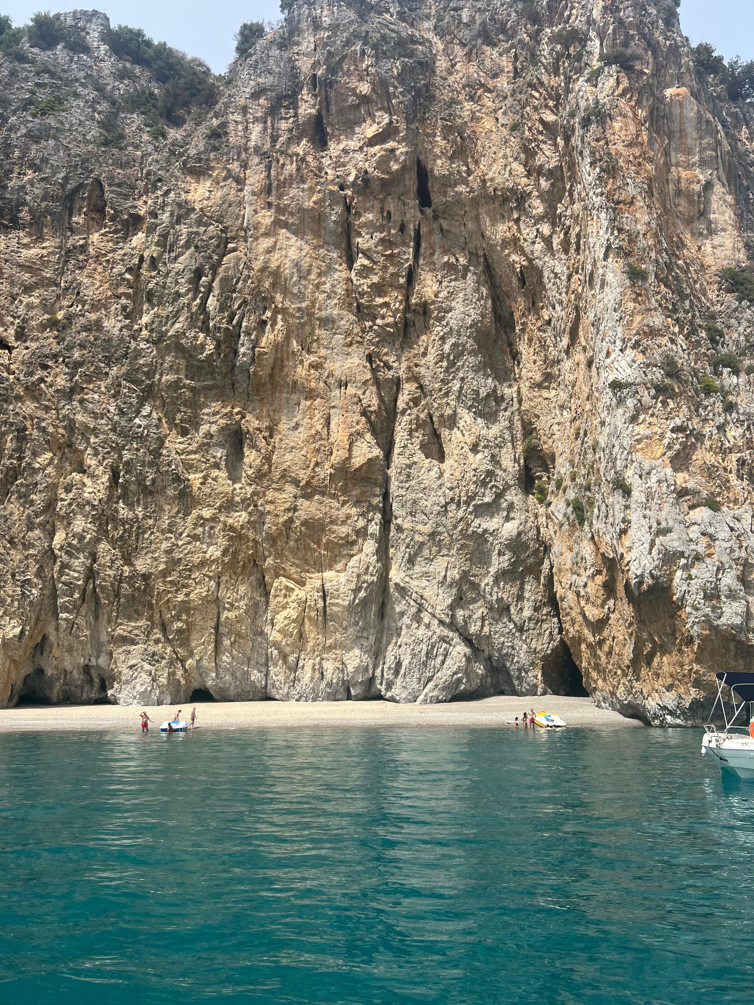 Scogliera rocciosa con una piccola spiaggia e alcune persone vicino al mare, acqua di colore azzurro intenso.
