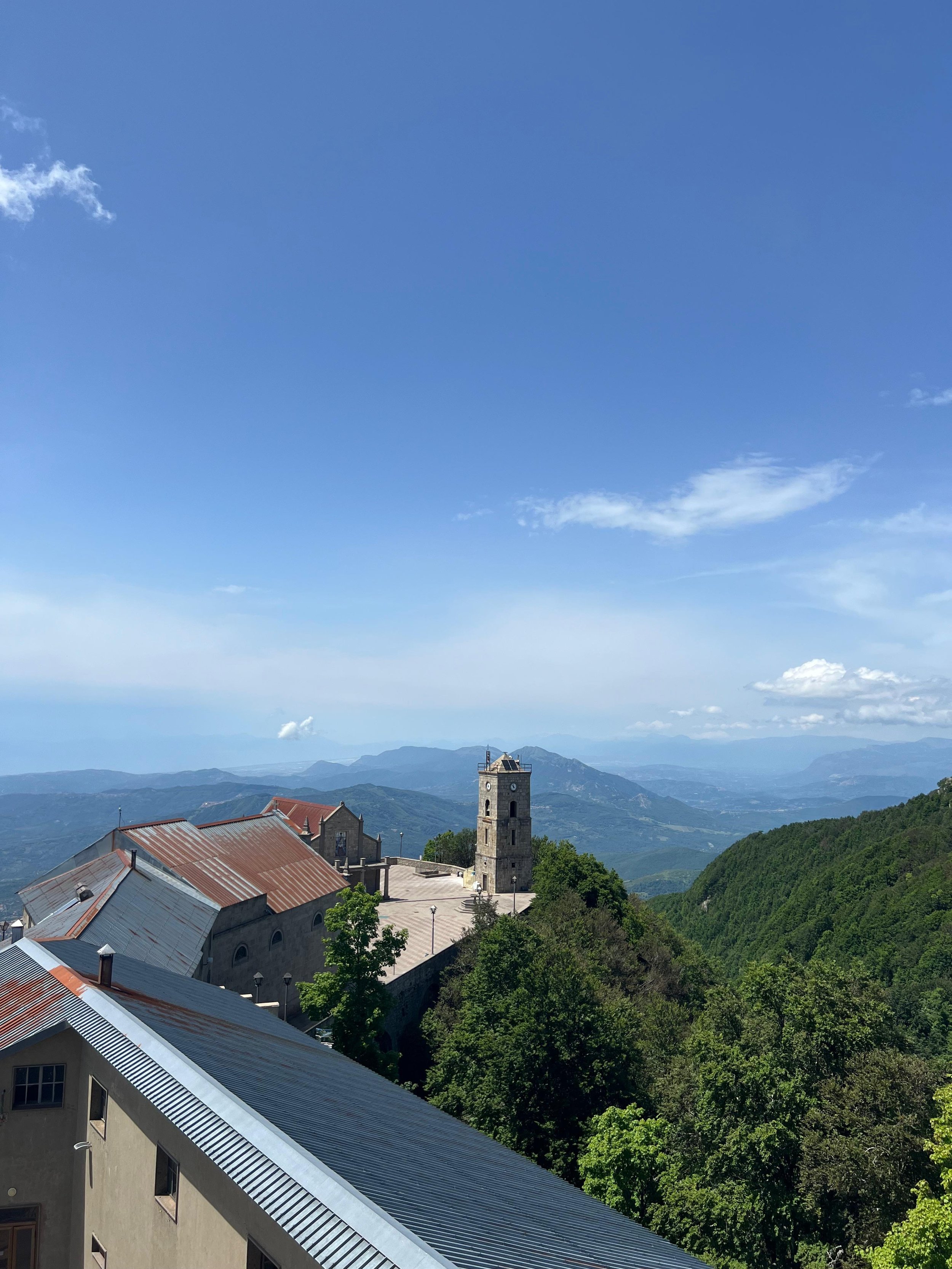 Vista di un monastero montano con un campanile in pietra, circondato da montagne e vegetazione, sotto un cielo sereno.