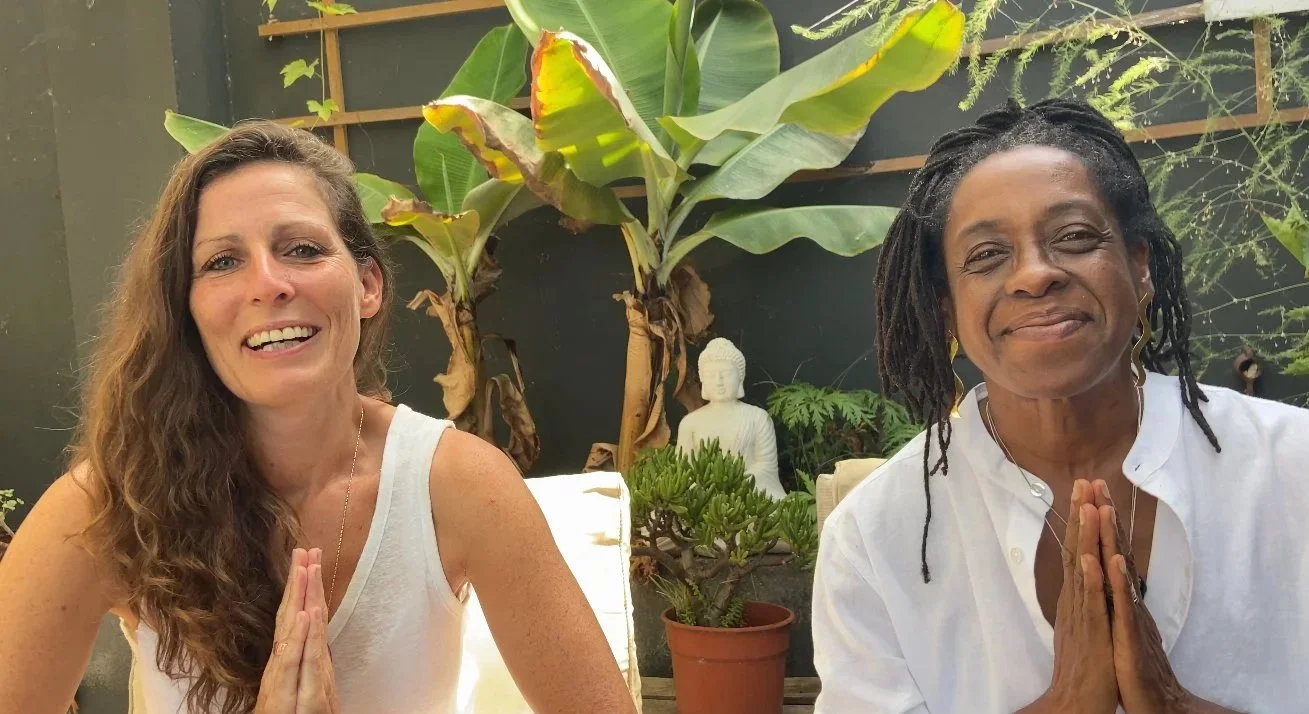 Two women practicing yoga or meditation with hands in prayer position, sitting outdoors with lush green plants and a small Buddha statue in the background.