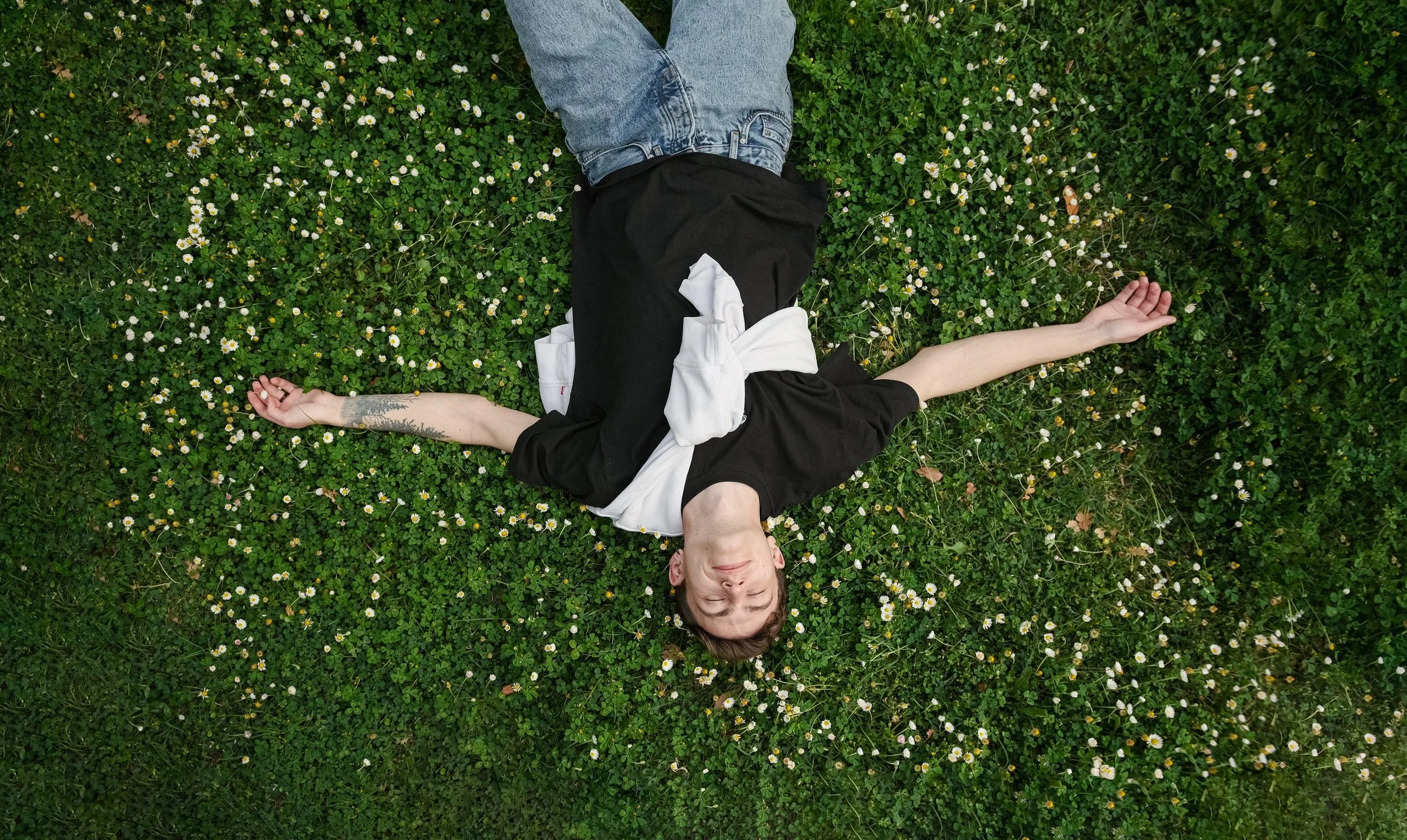 Person lying on a bed of green grass and small white flowers with closed eyes, wearing a black shirt and jeans, with arms stretched out and a white cloth draped over their chest.