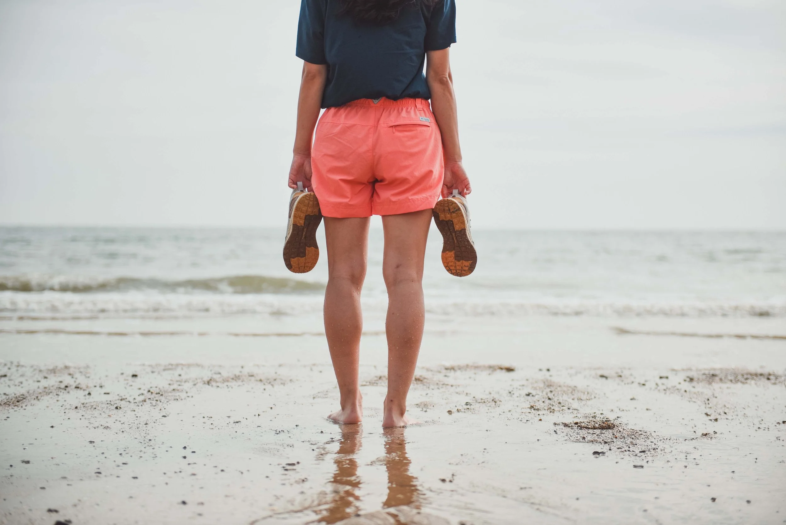 Person standing on a beach holding shoes, wearing pink shorts and a dark shirt, with the ocean and cloudy sky in the background.