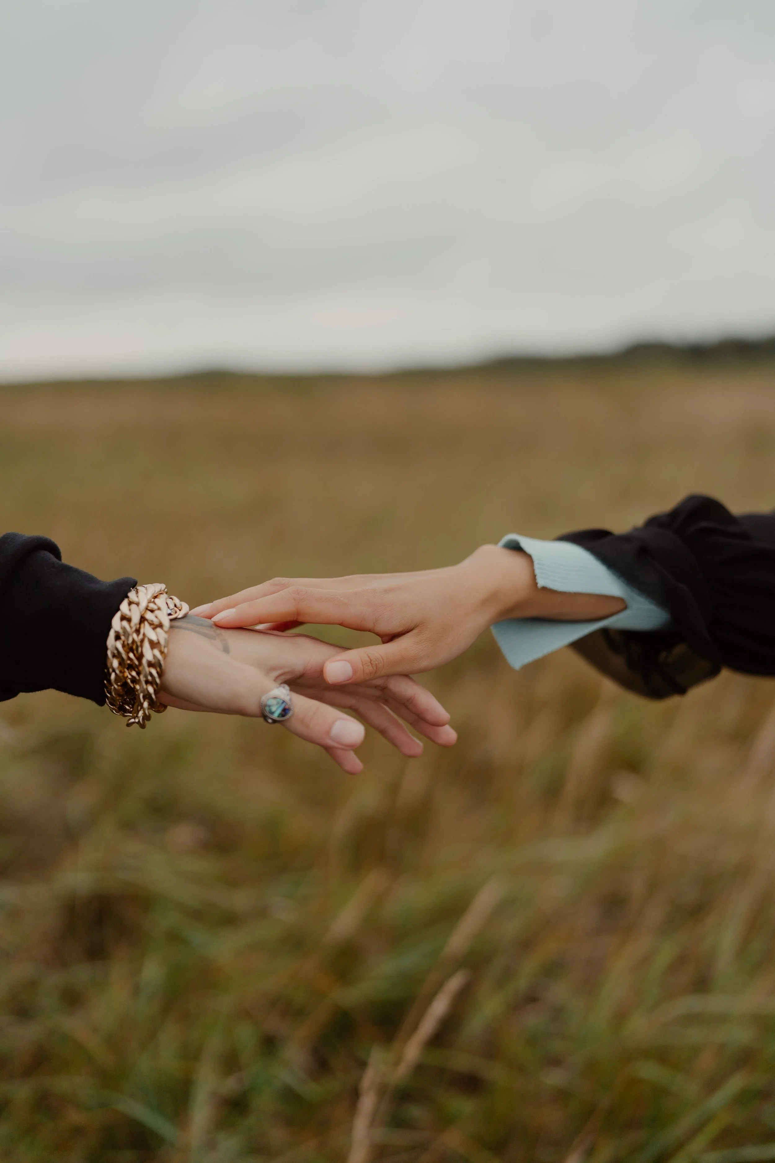 Two people reaching their hands to each other in an open grassy field on a cloudy day.