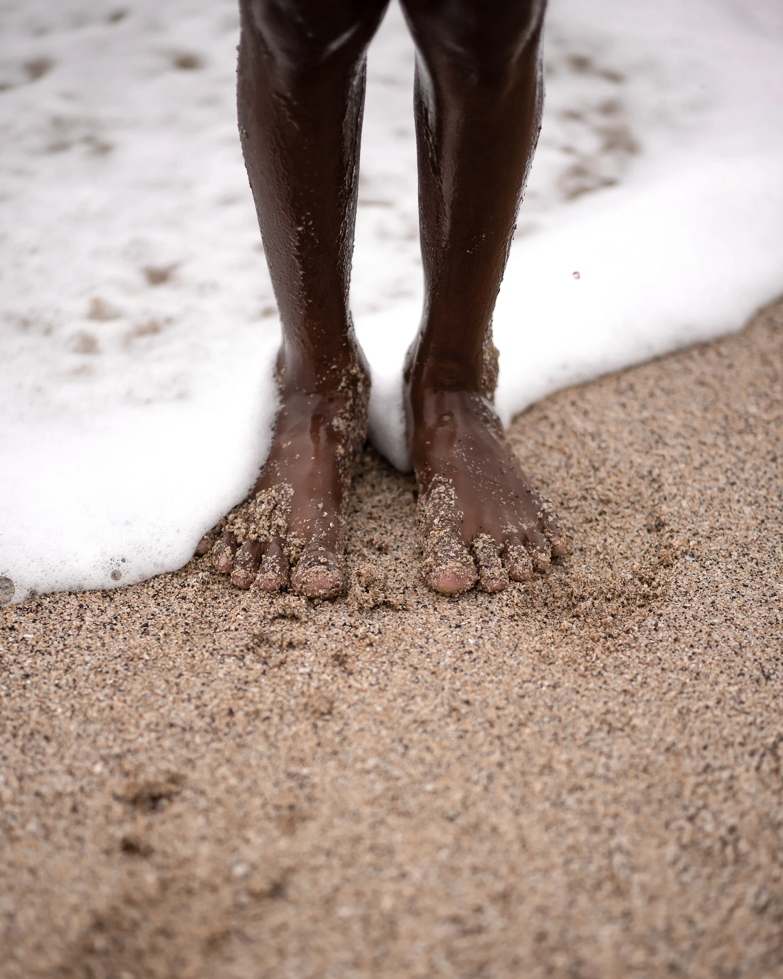 Close-up of a person's dark-skinned legs and feet standing on a sandy beach with foam from a wave nearby.