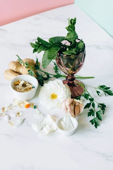 Assorted herbs and plants, including mint, ginger, garlic, and flowers, arranged on a white surface with a pink and mint background.