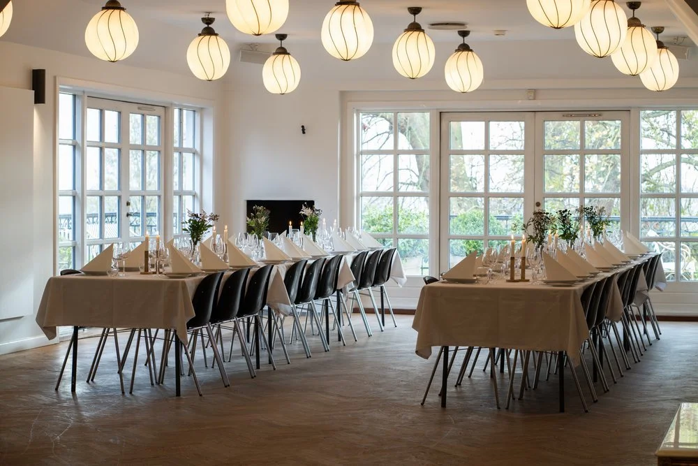 Festlokale med bordopdækning - Stilfuldt festlokale med dækkede borde og store vinduer, der giver naturligt lys.