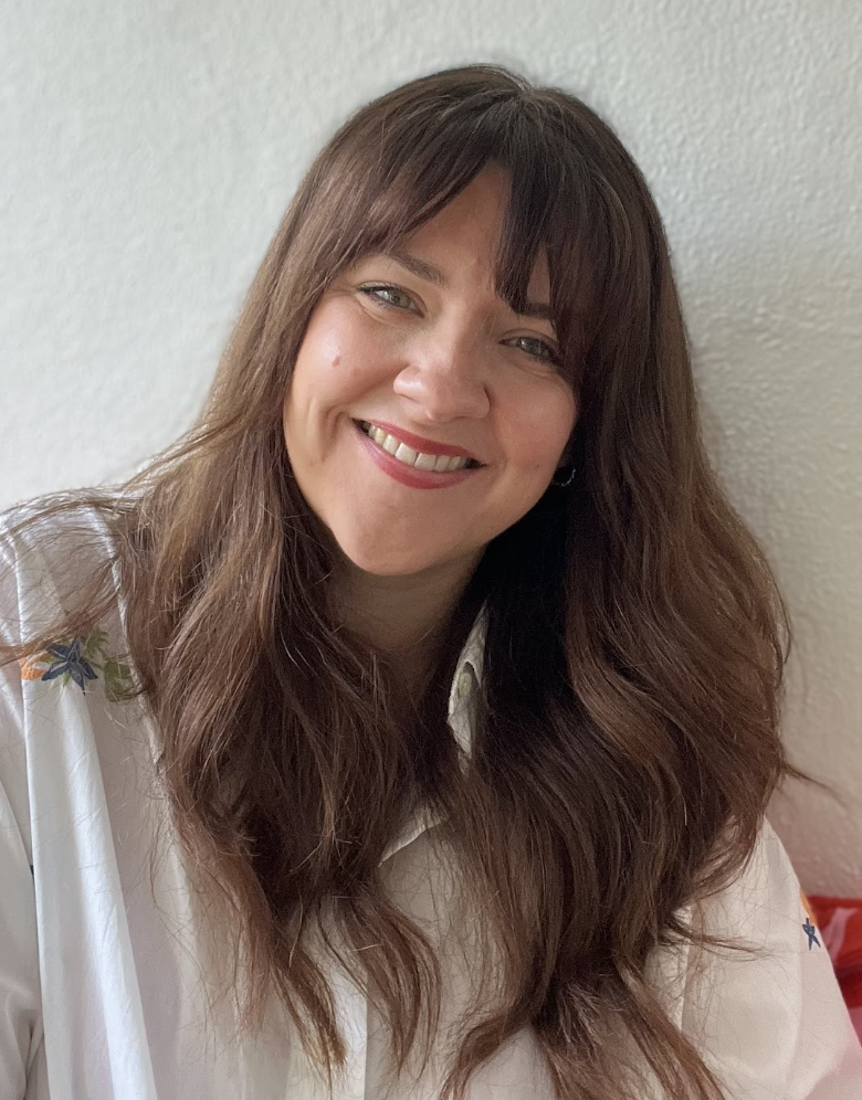 A woman with long, wavy brown hair and bangs smiling at the camera, wearing a white shirt with floral embroidery on the shoulder, standing against a textured light-colored wall.