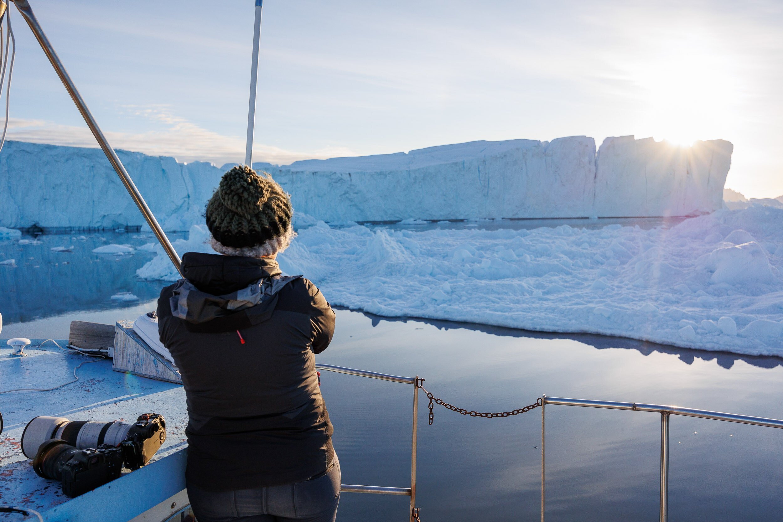 Person in winter jacket and knit hat on boat, looking at iceberg and calm water in icy landscape at sunset.