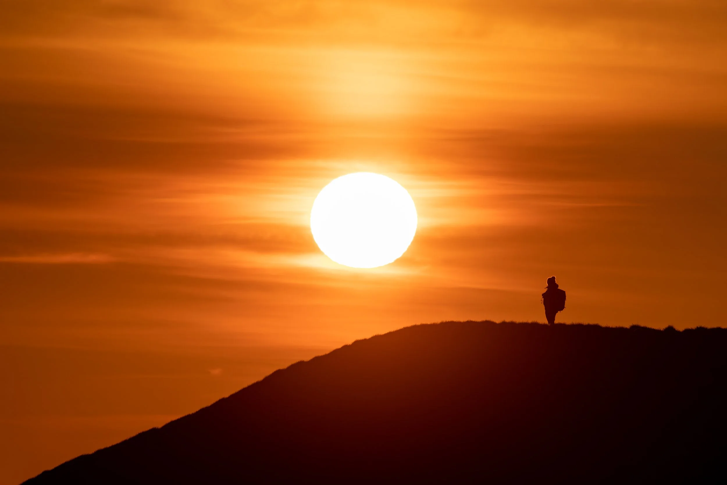 Silhouette of a person standing on a hill during sunset or sunrise, with a large, bright sun in the sky and orange clouds and sky in the background.