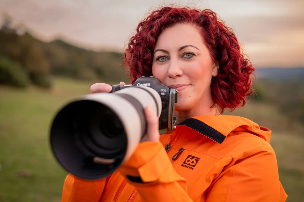 Woman with red curly hair holding a camera outdoors in a scenic landscape during sunset, wearing an orange jacket.