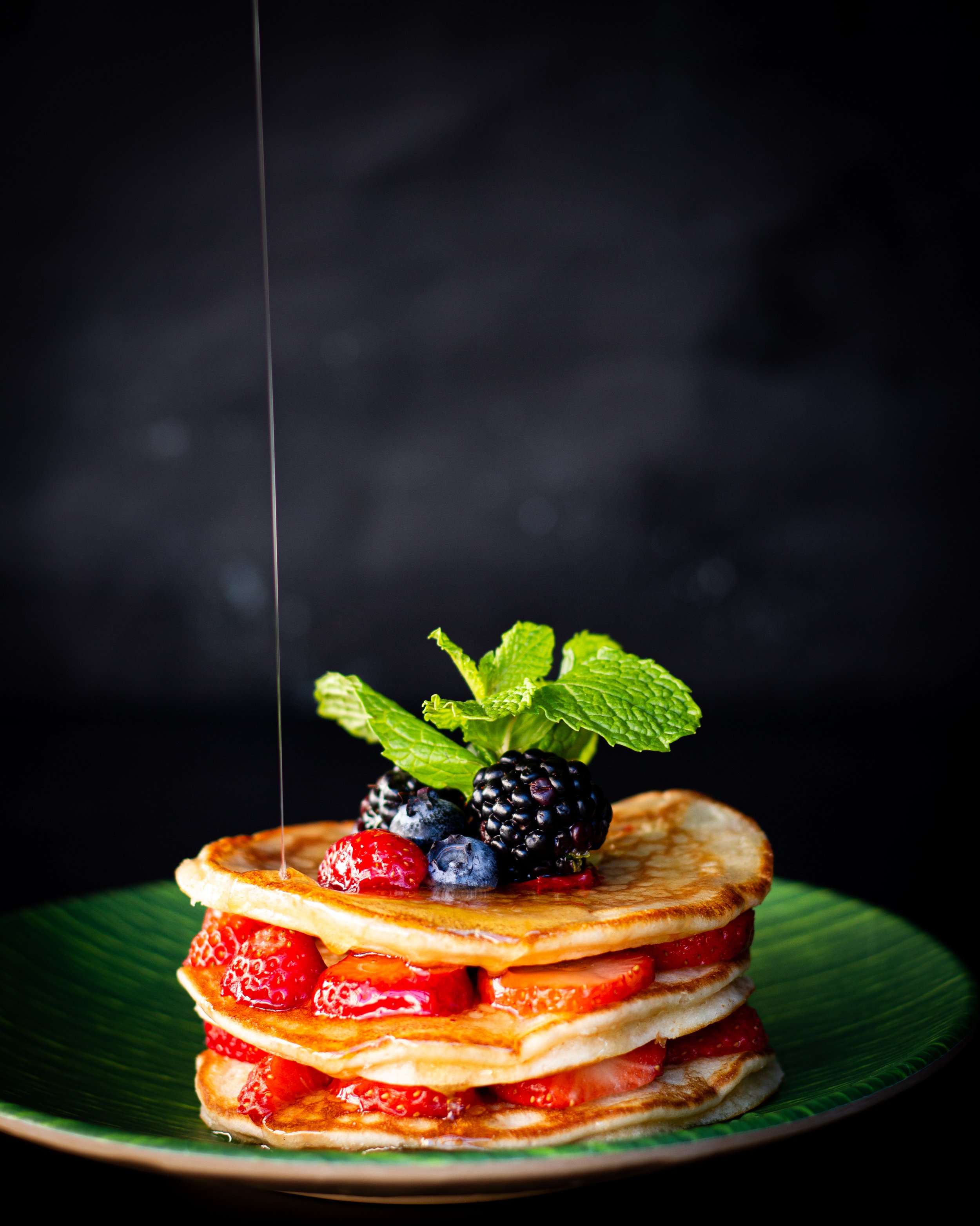 Una torre de pancakes dorados y esponjosos se alza sobre un plato verde oscuro, cada uno intercalado con rodajas brillantes de fresa y adornado con un montón de moras y arándanos jugosos en la cima. Un chorro de miel ámbar cae en cascada sobre la pil