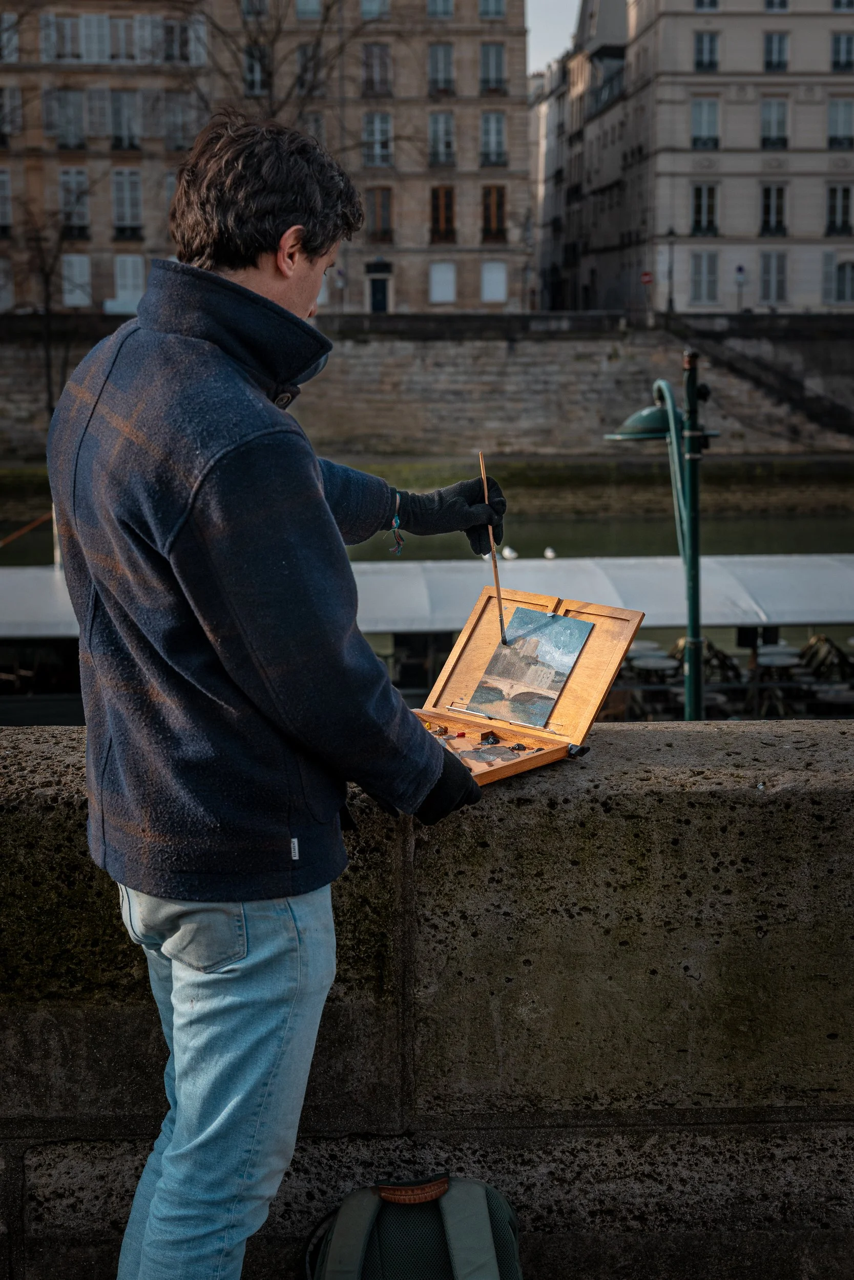 A man in a dark jacket paints on a small easel by a river with historic buildings in the background.