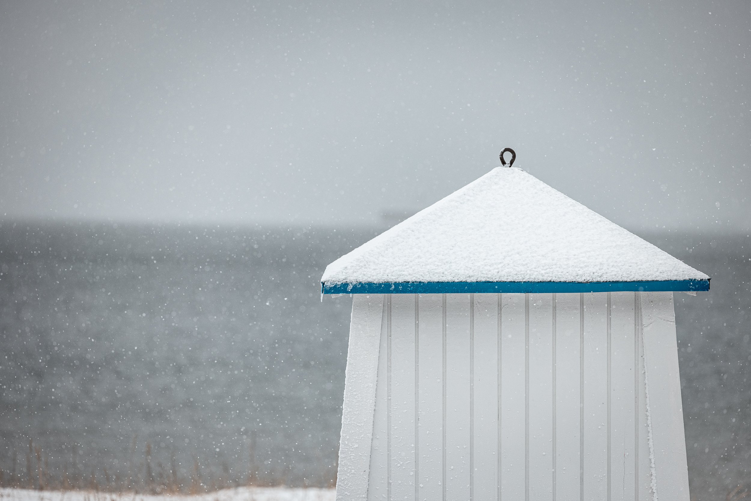 A snow-covered small structure or shed with a blue edge, set against a background of a snowing landscape and a blurred body of water.