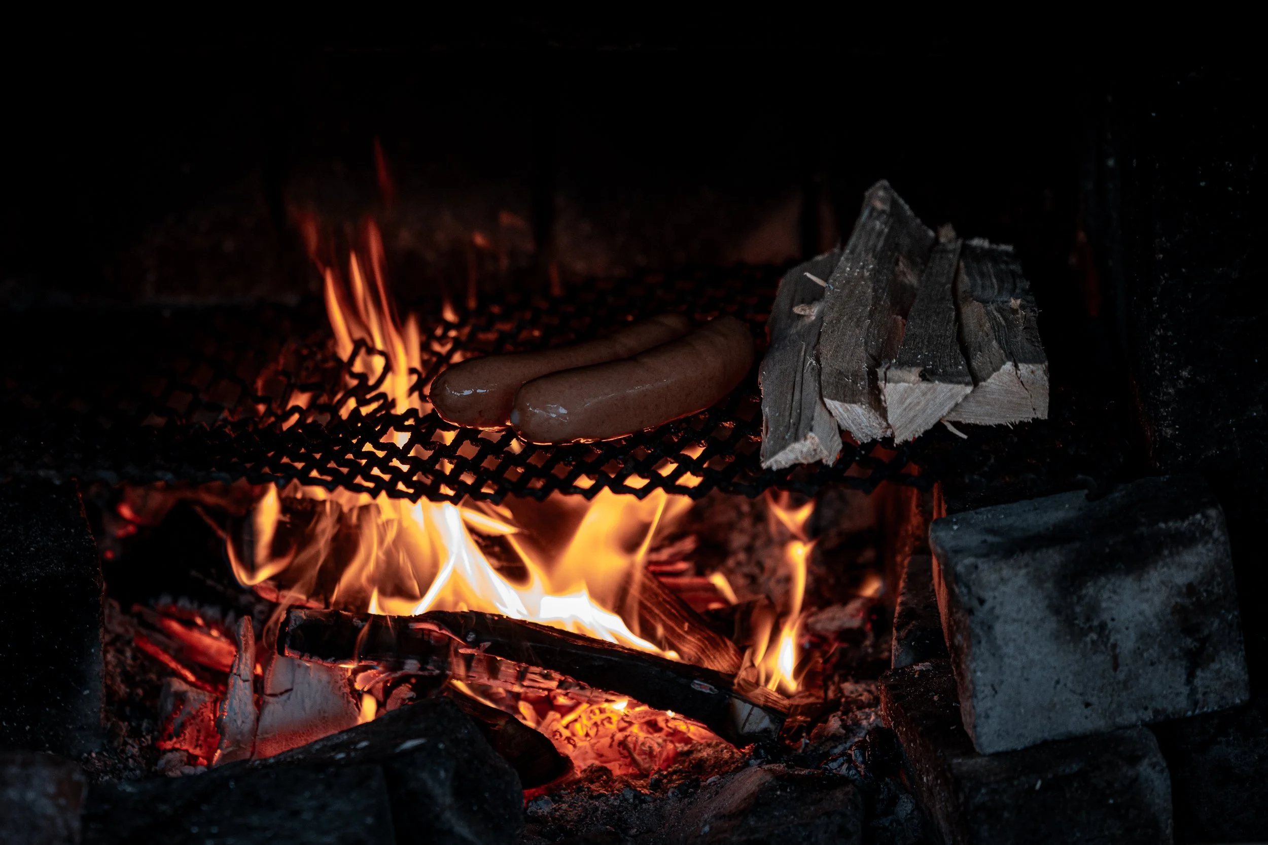 Hot dogs roasting over an open fire with firewood beside them.