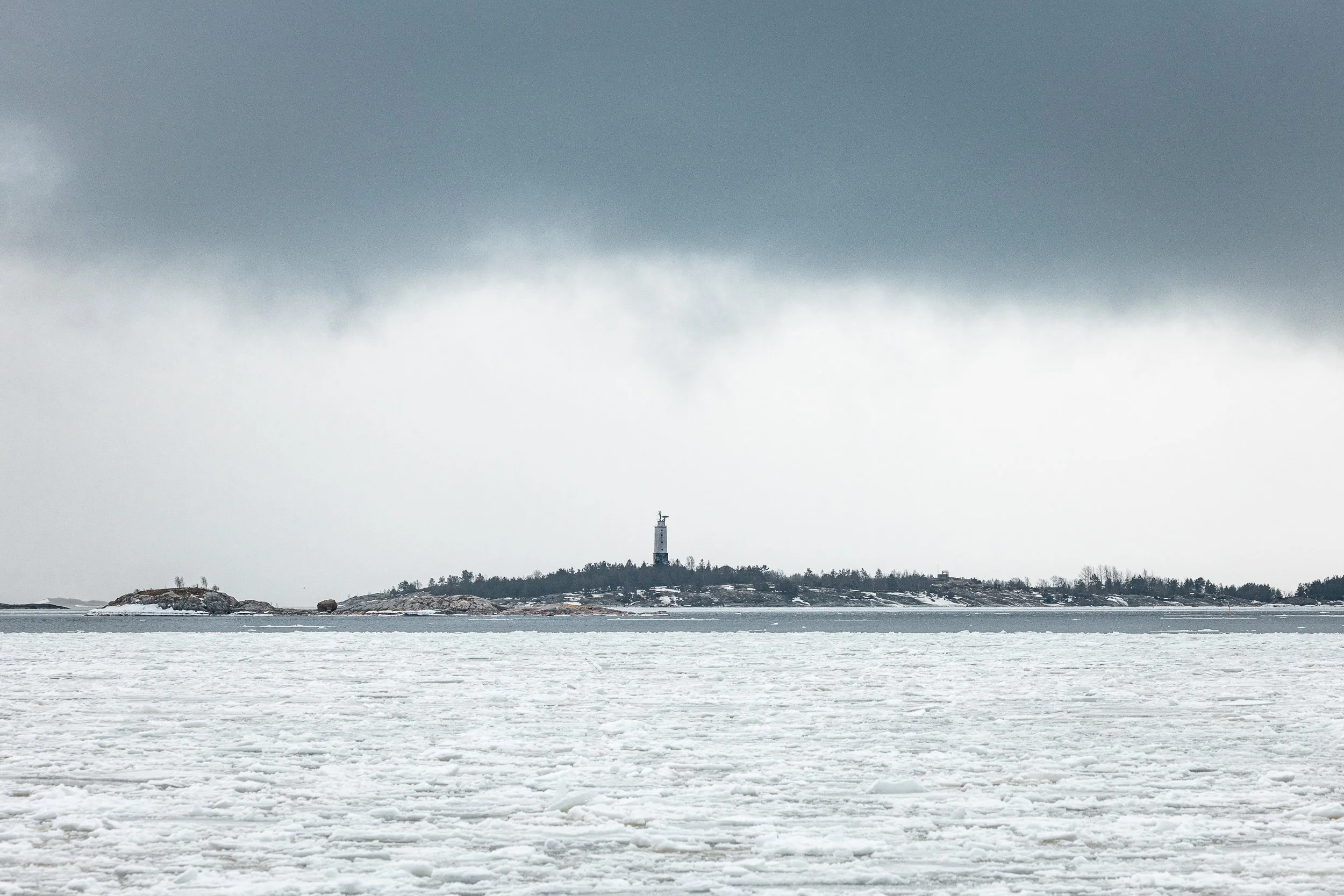 Snow-covered icy landscape with a distant island topped by a tall lighthouse and sparse trees, under a cloudy overcast sky.