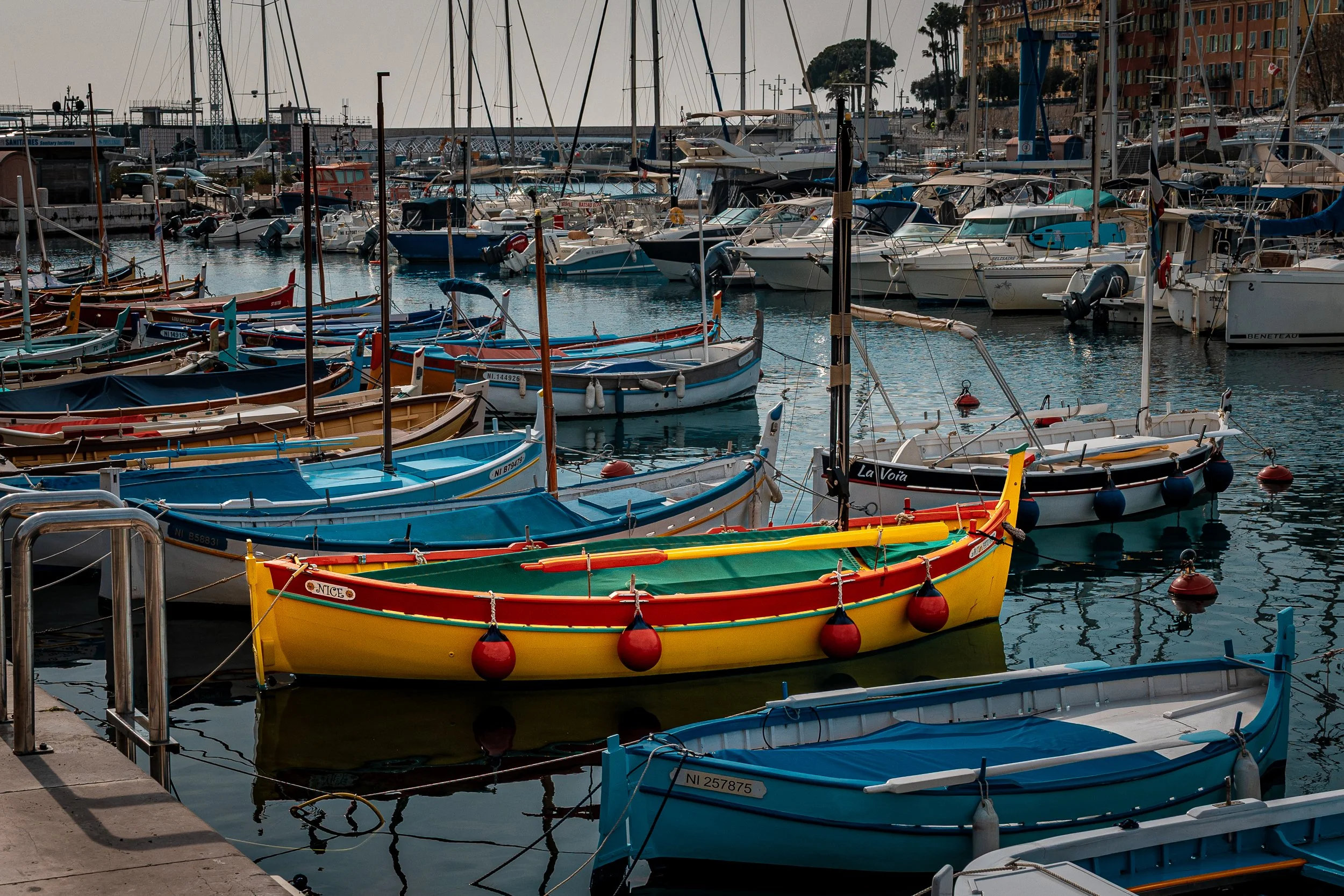Colorful small boats docked at a marina with larger yachts in the background, some buildings and trees are visible along the shoreline.