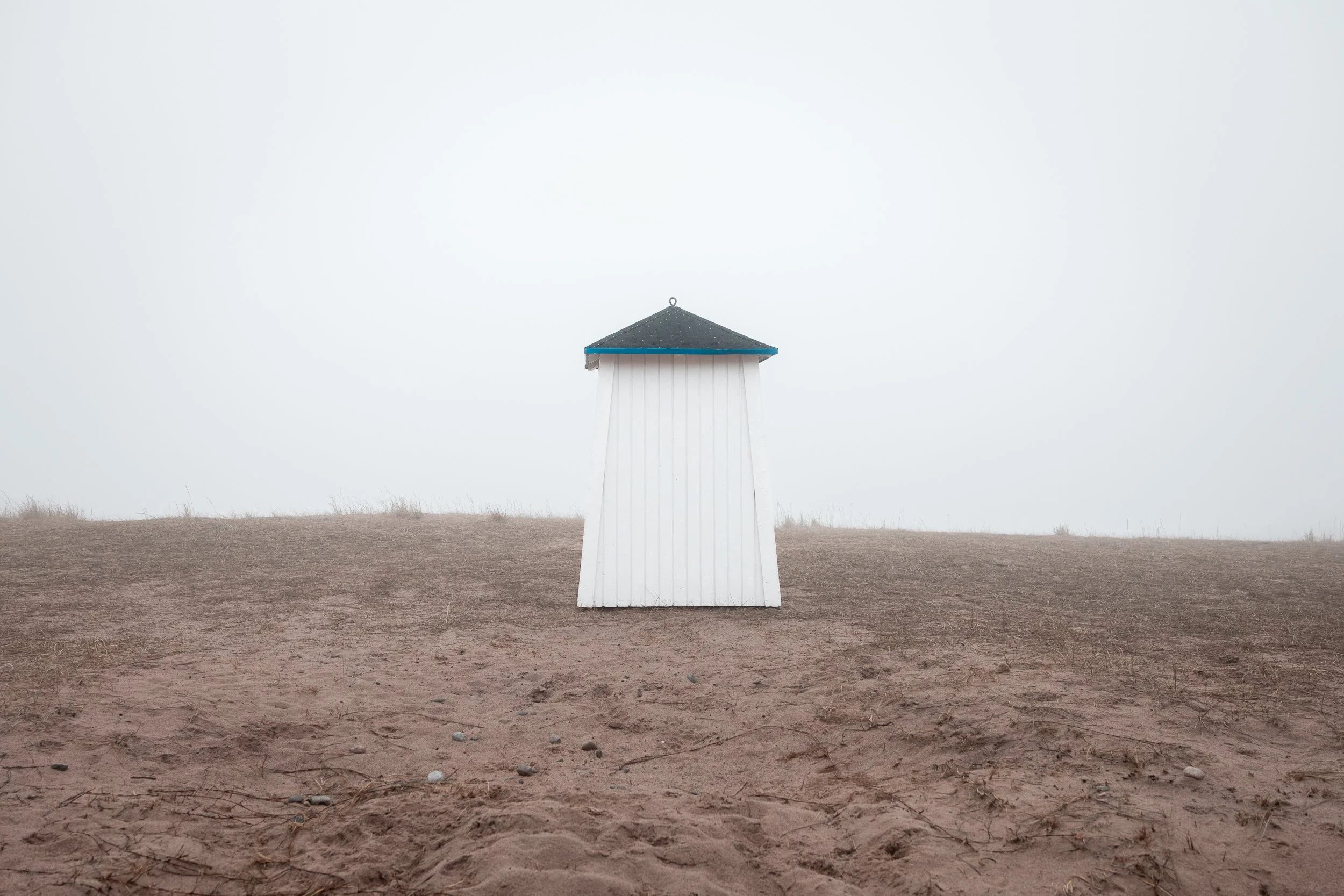 A small white structure with a dark sloped roof stands alone on a sandy, barren landscape under a foggy sky.