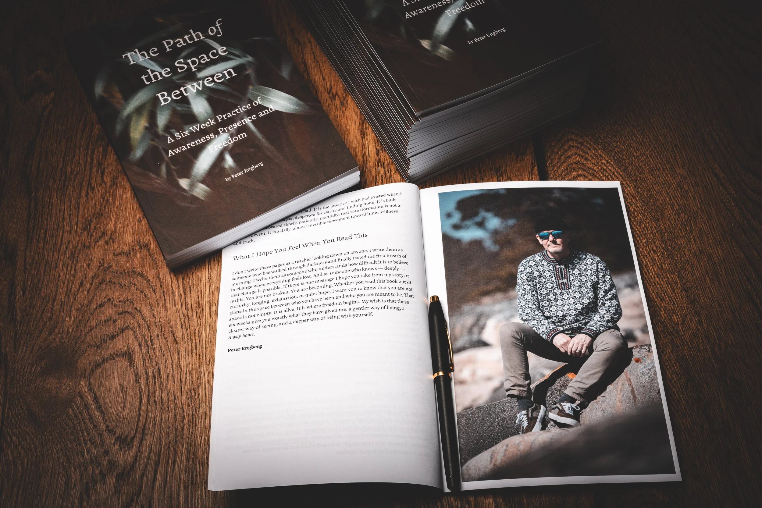 Open book with a photograph of a man sitting on rocks on right page, and text on the left page. Also, a closed book titled 'The Path of the Space Between' and a stack of similar books on a wooden surface.
