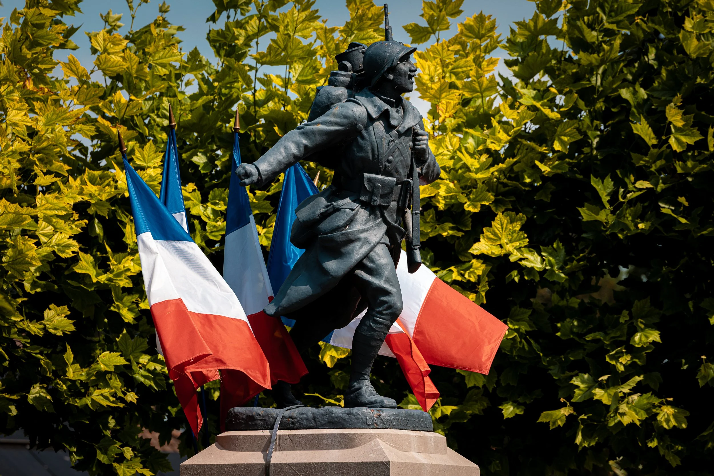 A bronze statue of a World War II soldier on a pedestal with French flags in the background, surrounded by green foliage.